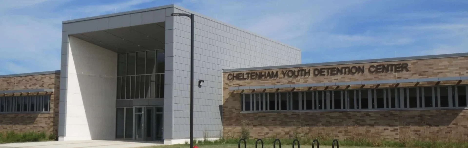 Exterior view of Cheltenham Youth Detention Center building with brick and white panel facade, glass entrance doors, a black streetlight, and a sign displaying its name.