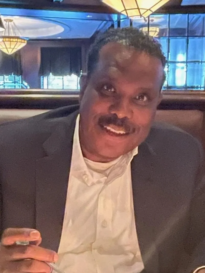 A man with a mustache smiling, wearing a dark sweater over a light blue shirt with a patterned tie, inside a modern office building with glass walls.