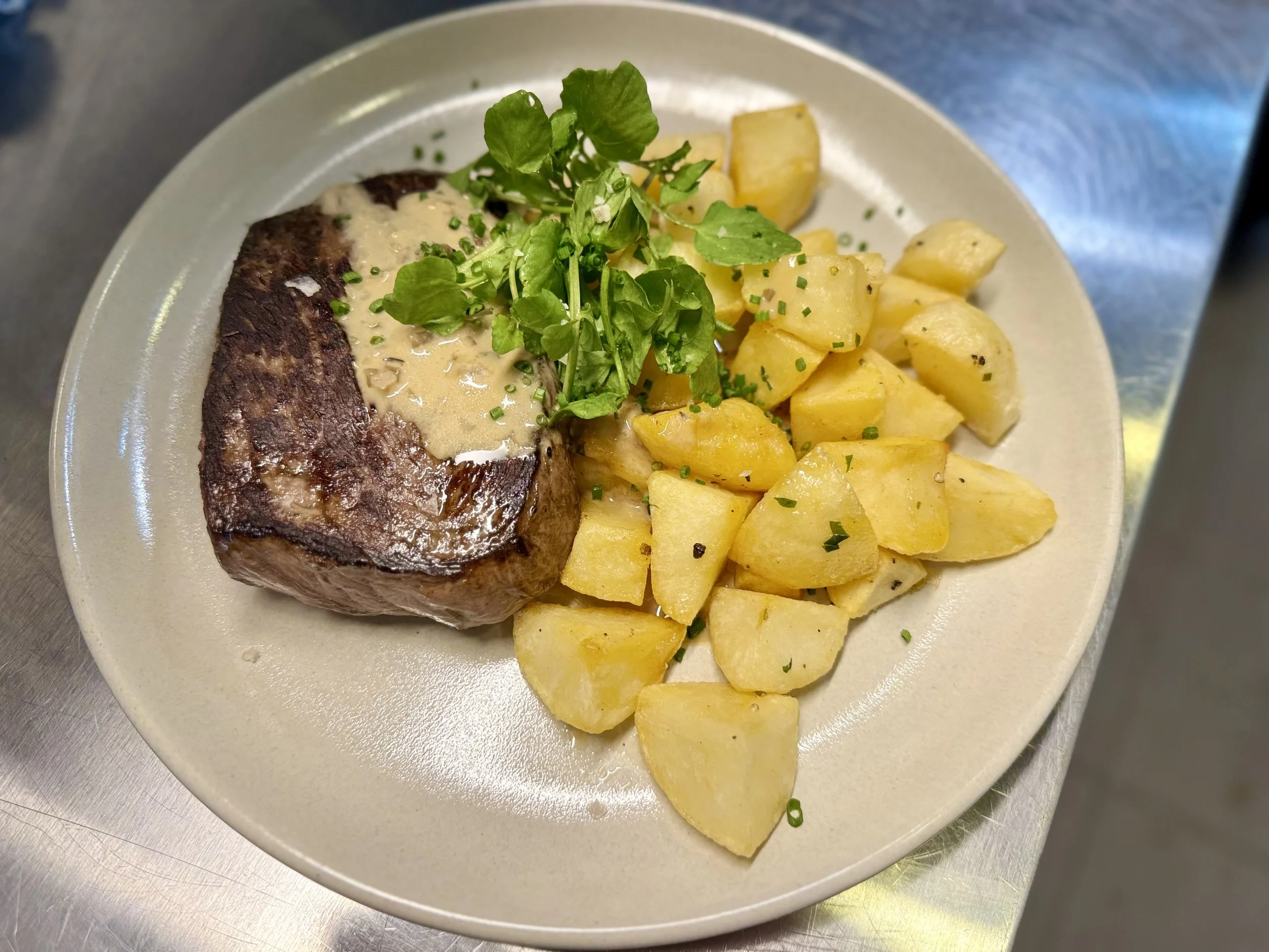 A plate with cooked steak topped with a creamy sauce, green garnish, and chopped chives, served with roasted potatoes and a sprig of fresh herbs.