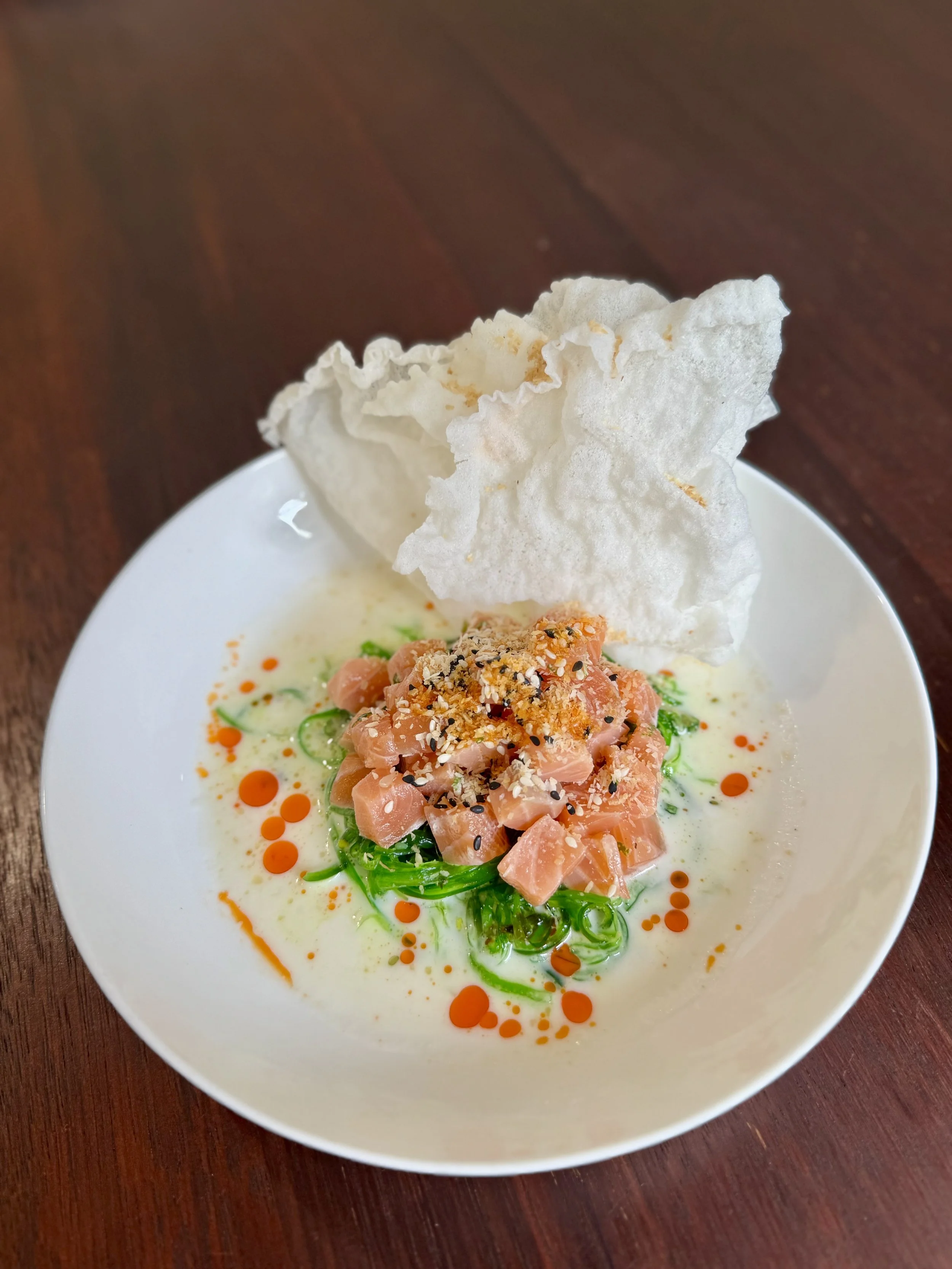 Dish of salmon tartare with green onion and sesame seeds, served with crispy rice crackers and drizzled with orange sauce in a white bowl on a wooden table.