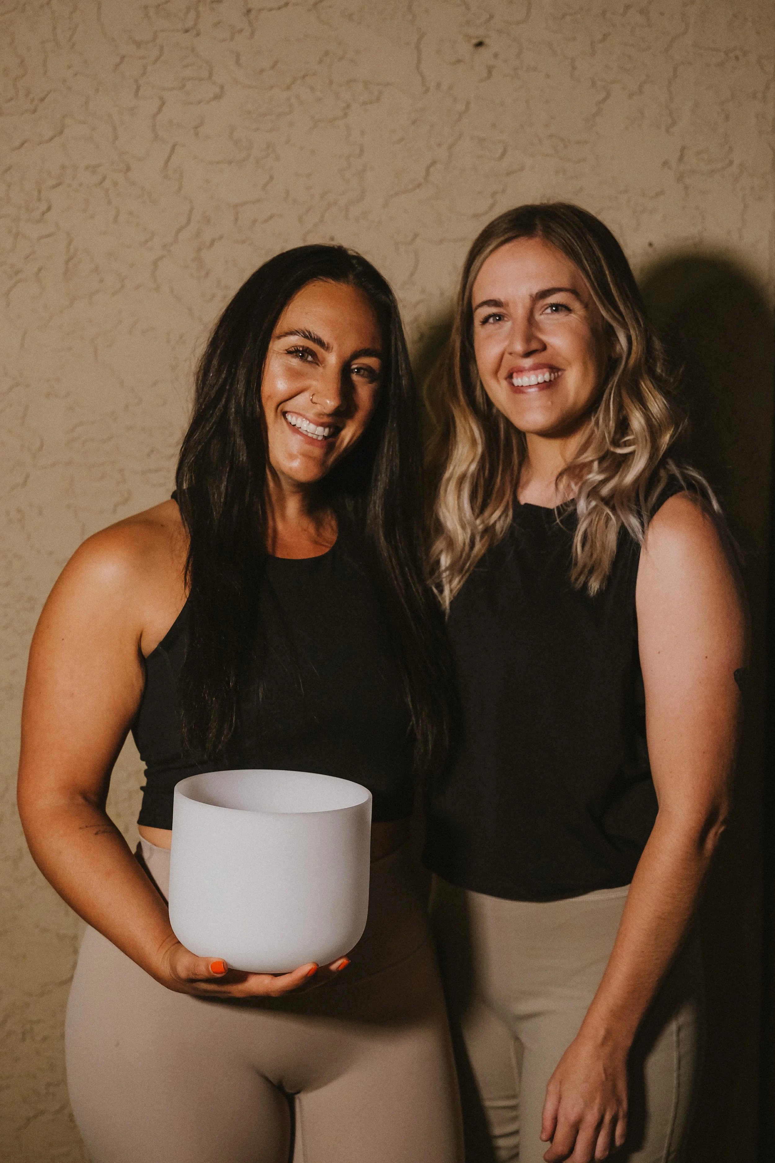 Two smiling women standing together against a beige textured wall, one holding a white singing bowl.