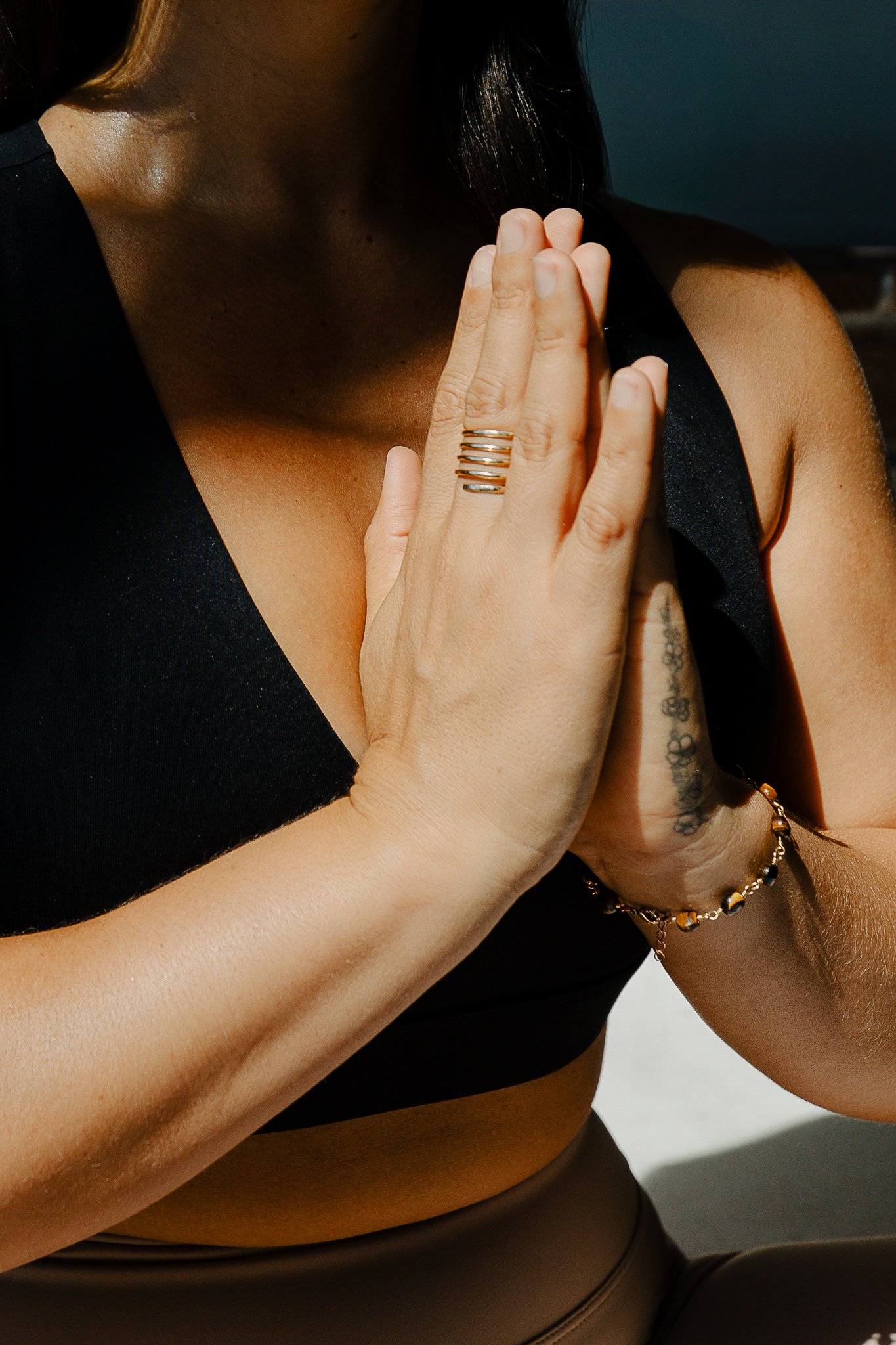 Close-up of a woman with tan skin wearing a black sports top and brown high-waisted pants, with her hands pressed together in a prayer or meditation pose, wearing rings and a bracelet, with sunlight highlighting her skin.