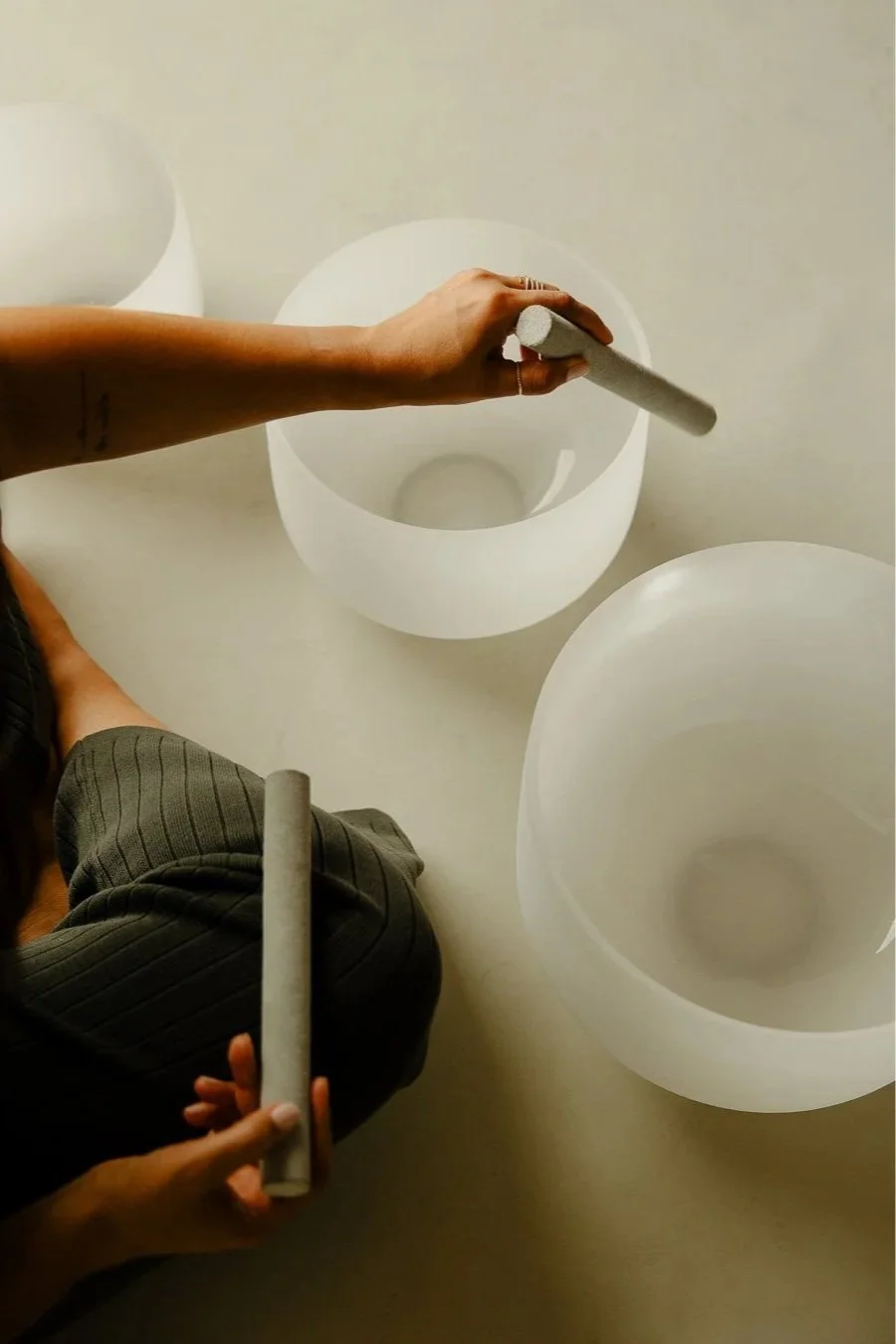 Person playing crystal singing bowls, captured from above, with one hand holding a mallet and two bowls on a table.