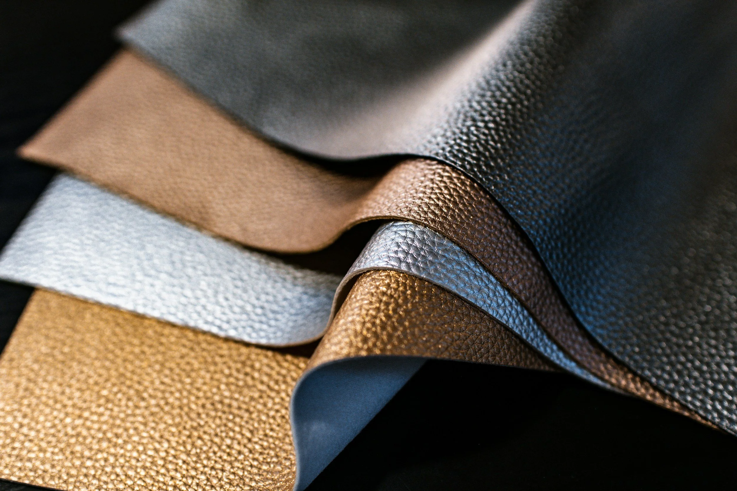 A close-up of textured leather fabric swatches in various metallic and neutral shades layered on top of each other.