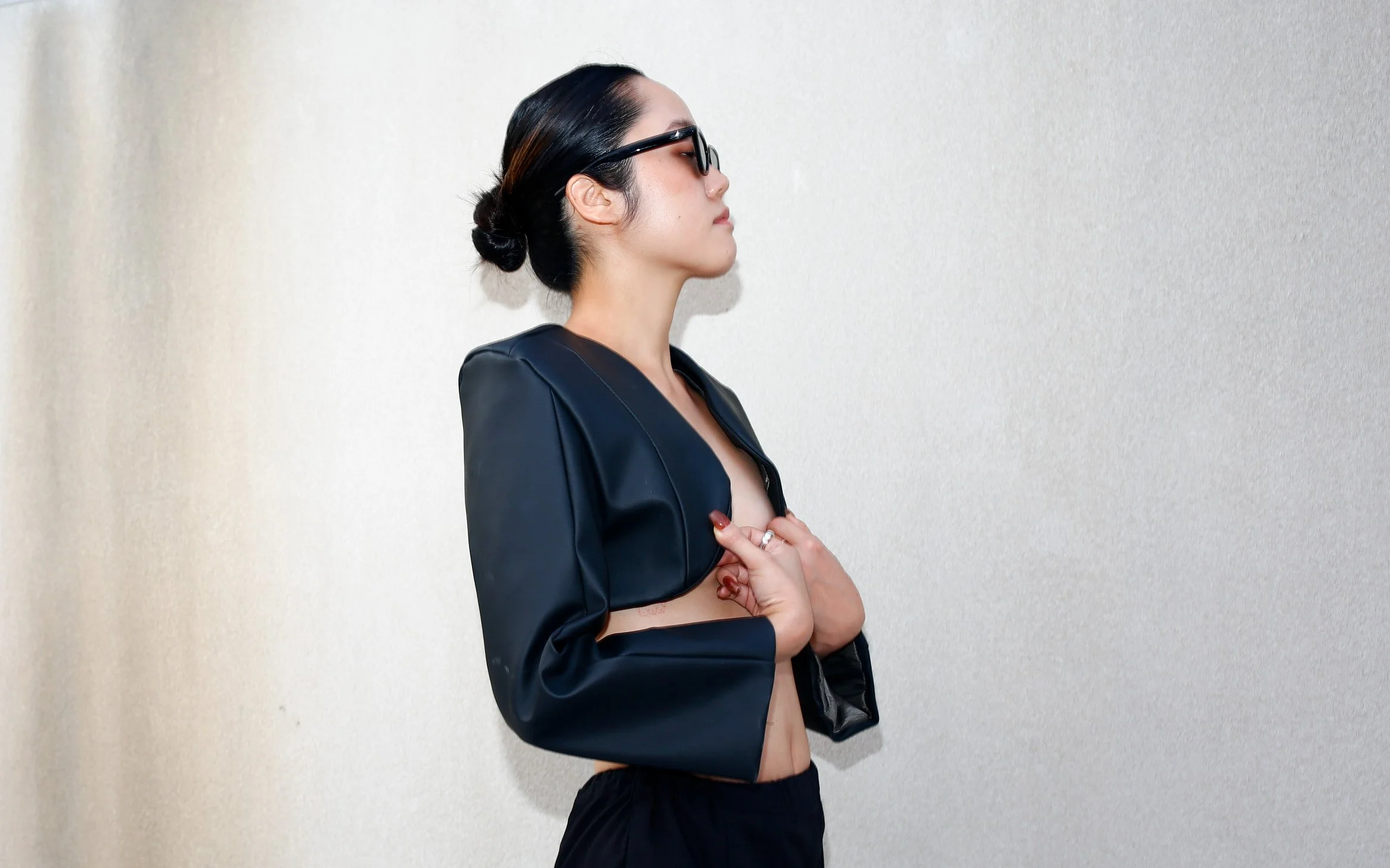 Side profile of a woman with dark hair in a bun, wearing black sunglasses, a black jacket, and black shorts, standing against a plain wall.