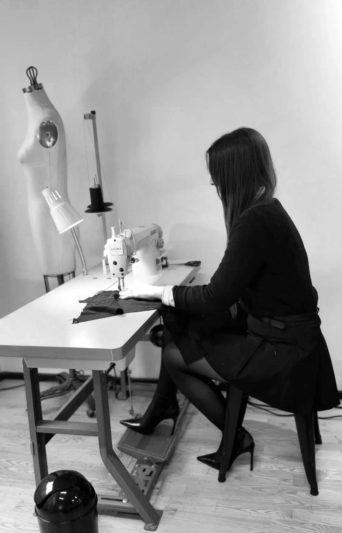 A woman sewing fabric on a sewing machine in a minimalistic room, with a dress form in the background and a small trash bin on the floor.