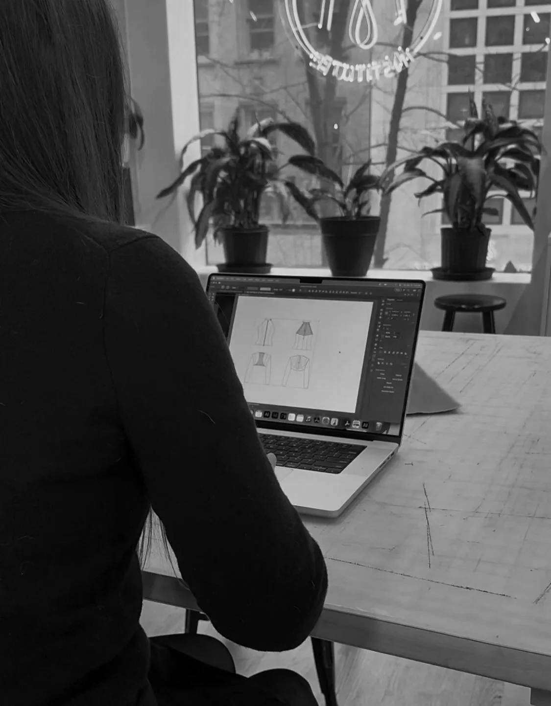 A woman working on a laptop at a wooden table in a room with large windows and potted plants, with buildings visible outside.