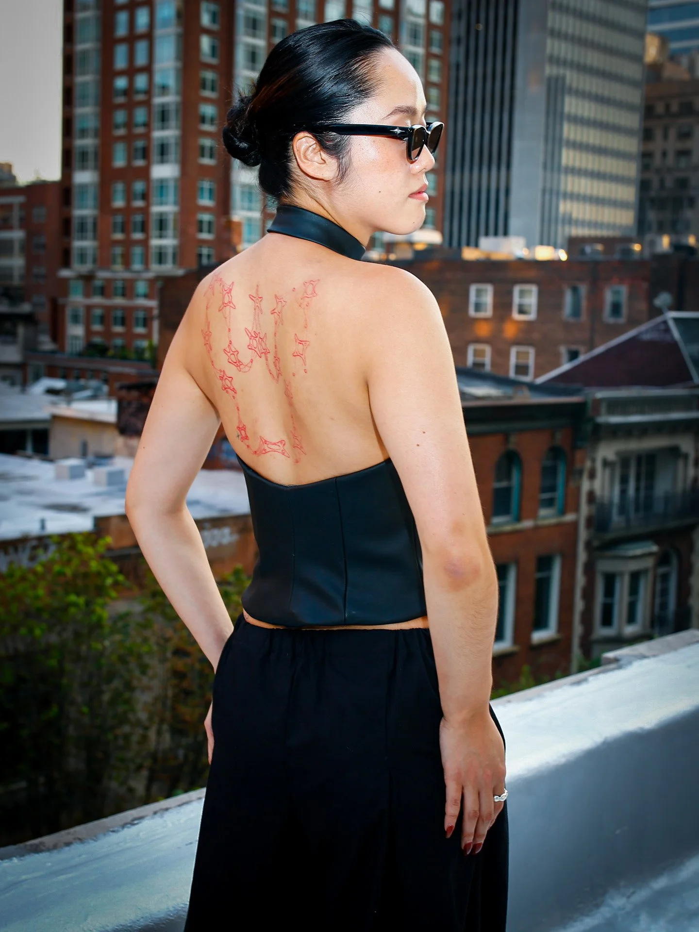 Woman with dark hair tied back in a bun, wearing sunglasses, black strapless top, and black pants, standing on a rooftop in an urban area with tall buildings in the background, showing a star tattoo on her back.