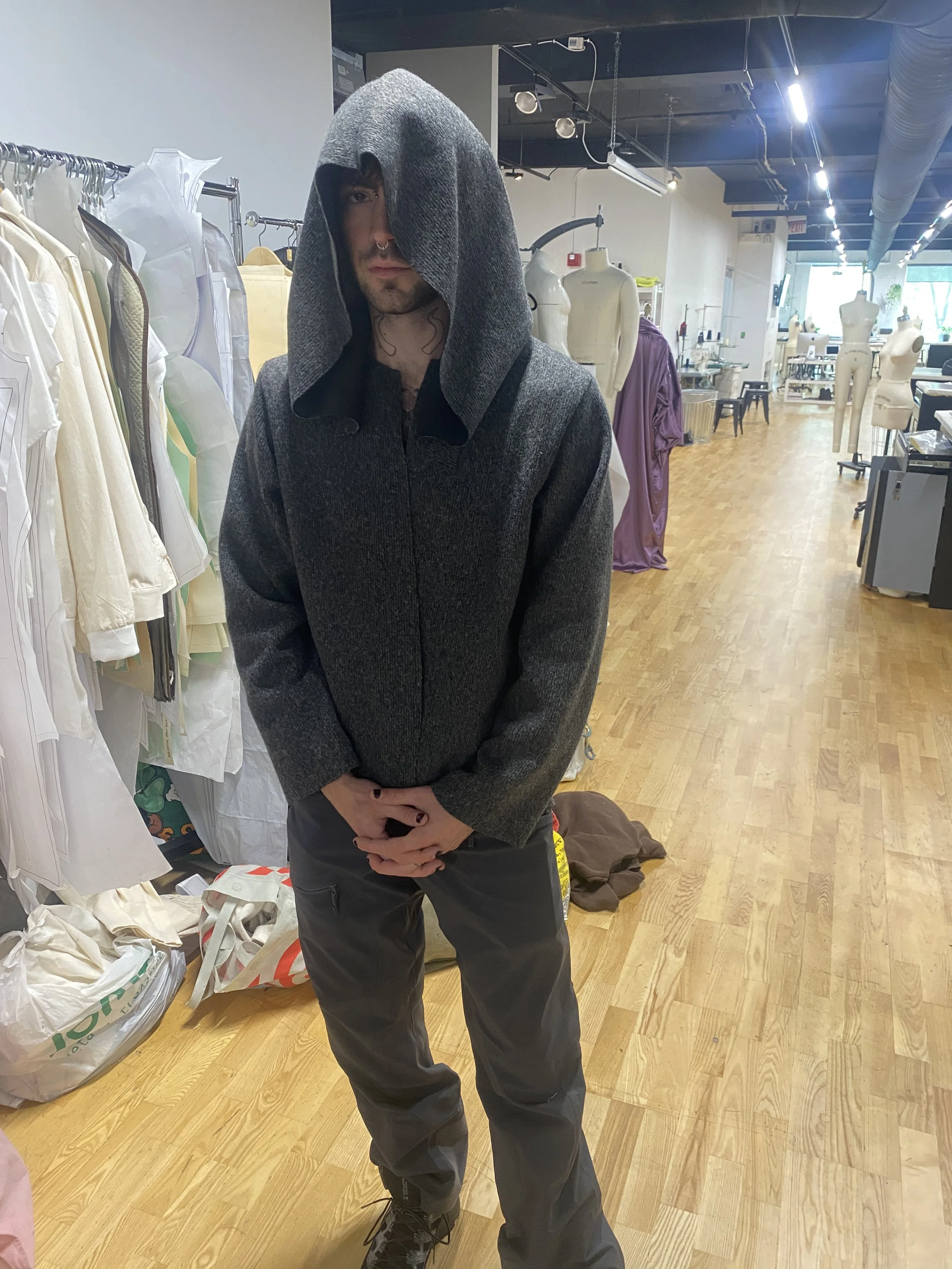 A person with dark hair, facial piercings, and dark clothing, standing indoors in a clothing store, with racks of clothing and mannequins in the background.