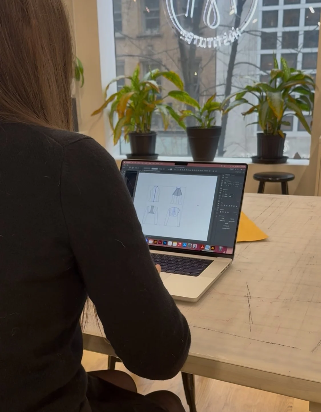 Woman working on a laptop at a wooden table, designing clothing sketches on screen, with potted plants and windows in the background.