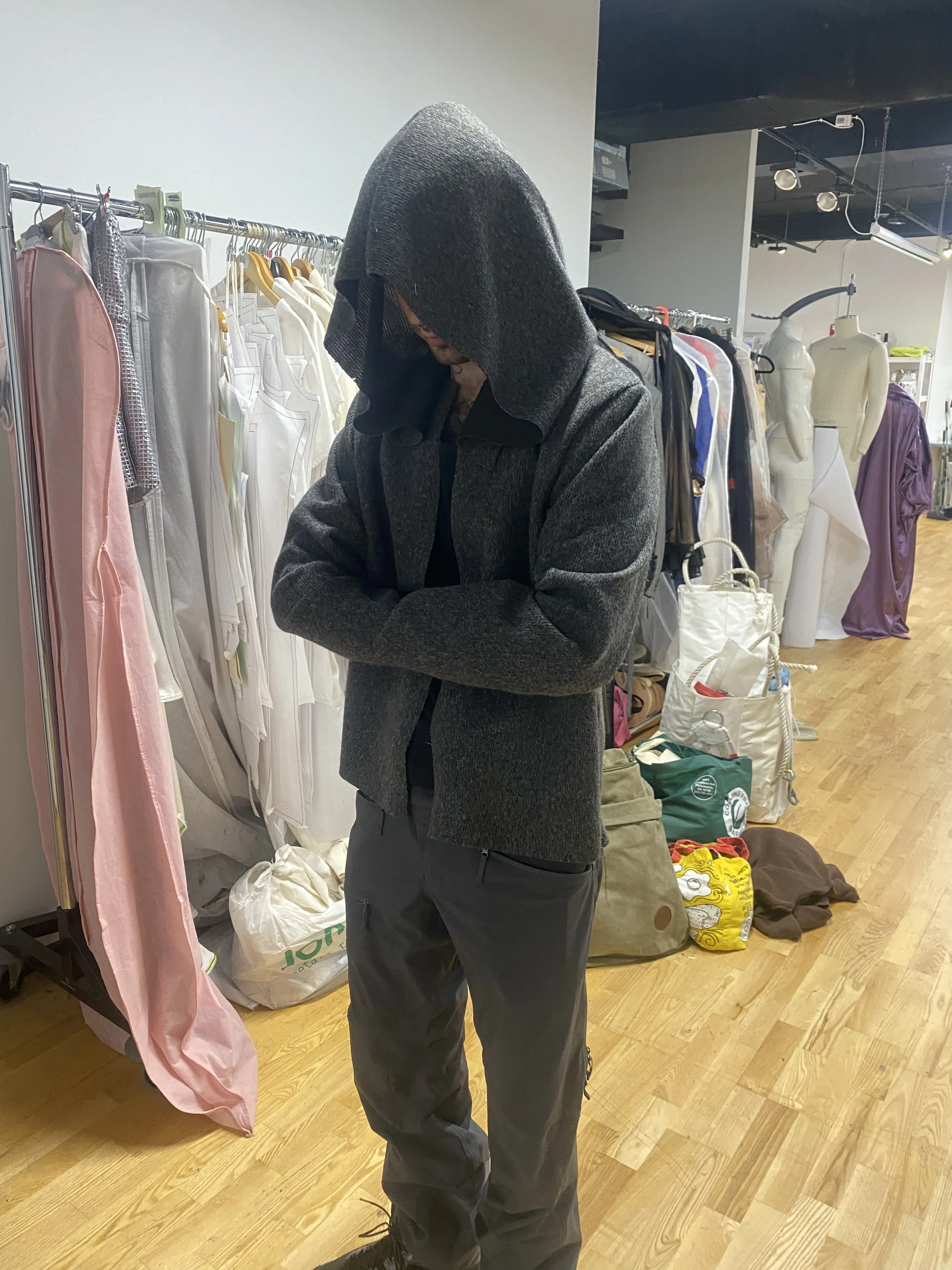 A person wearing a dark hoodie and jacket, standing inside a clothing store with racks of clothes and mannequins in the background.