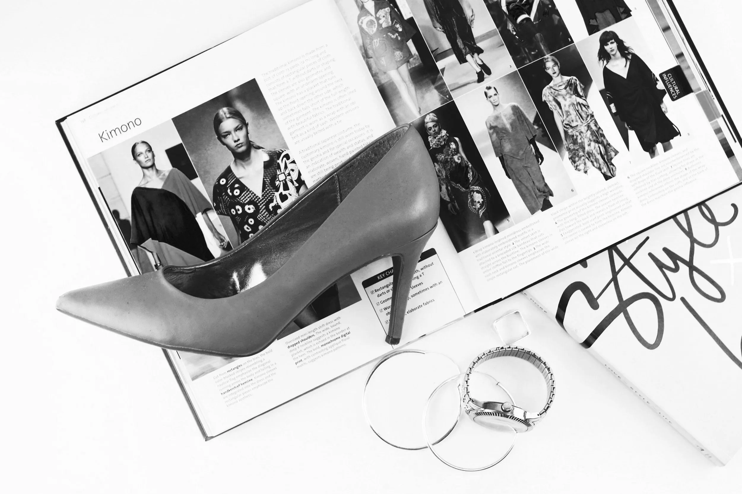 Black and white photo of a magazine spread open to pages with fashion models. In front of the magazine are a high heel shoe, a wristwatch, some bangles, and a pen on a white surface.