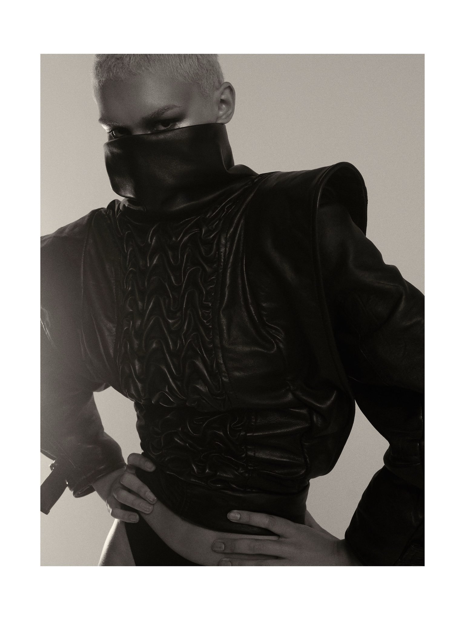 A person wearing a black face mask and a shiny black leather jacket, looking directly at the camera, with one hand resting on their waist and the other hand on their stomach.