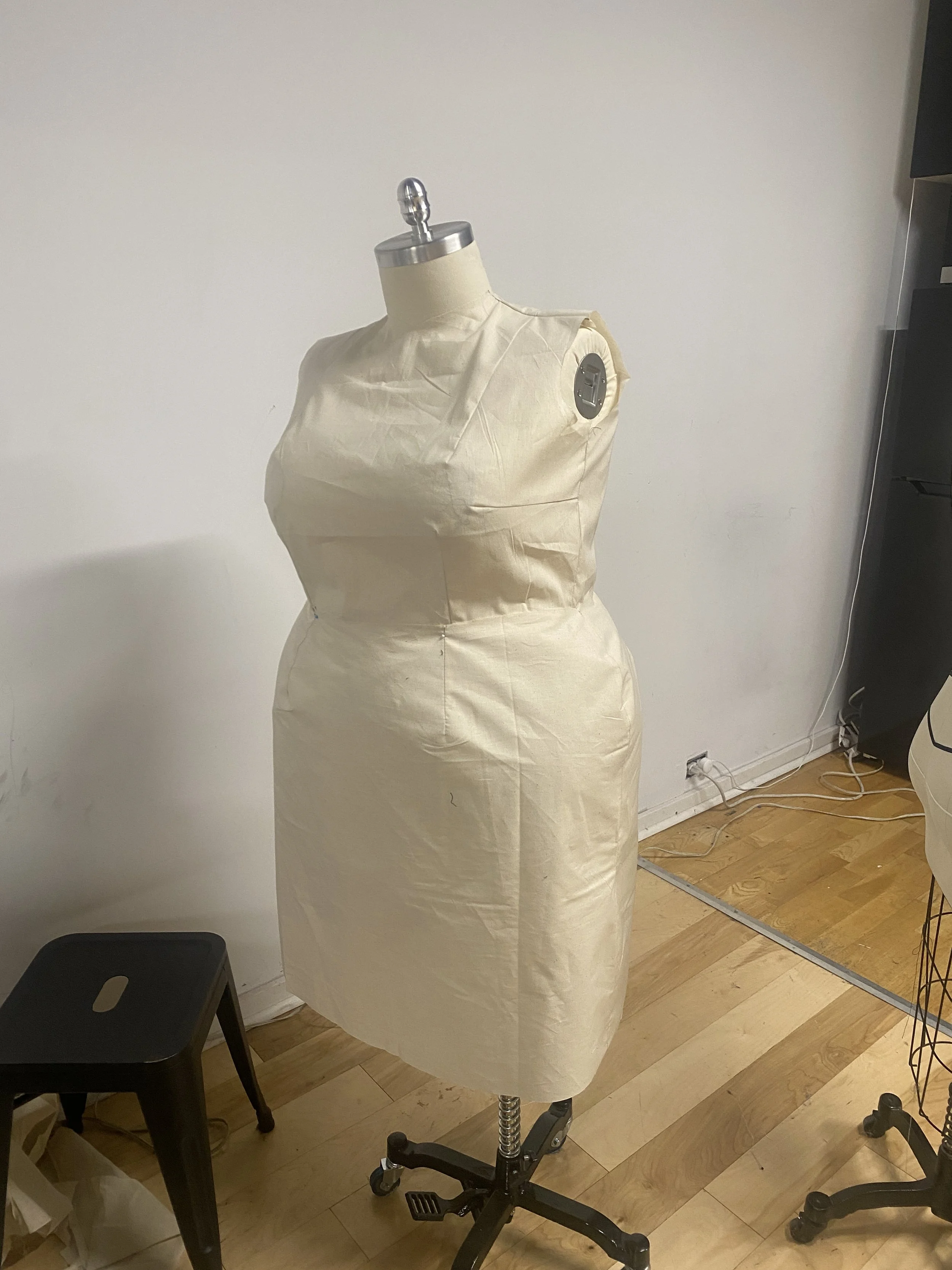 A dress form mannequin covered in beige fabric with an adjustable metal neck cap, set on a black wheeled stand in a room with wooden flooring, a black stool, and nearby electrical cords and furniture.