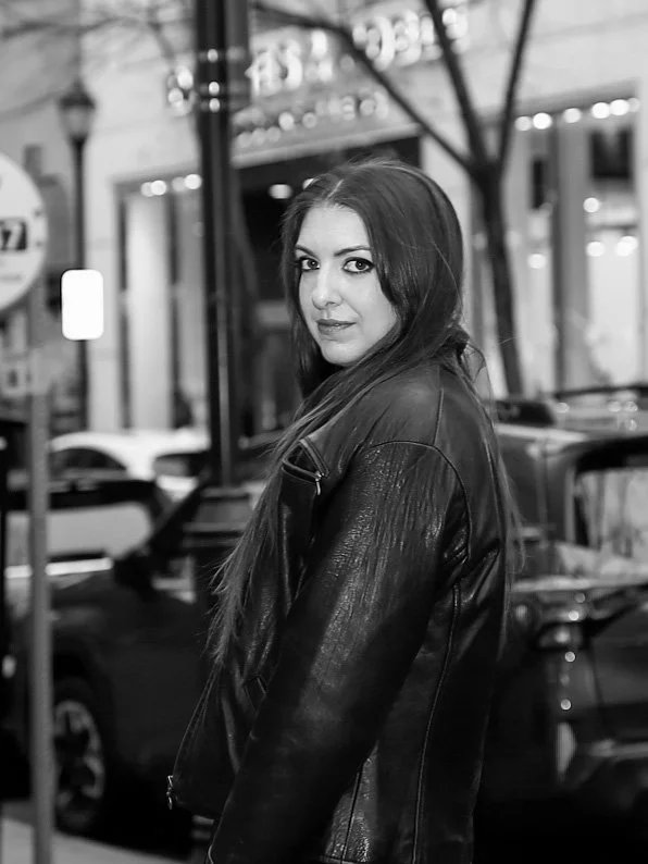 A woman with long hair wearing a leather jacket, standing on a city street at night, with cars and storefronts in the background.