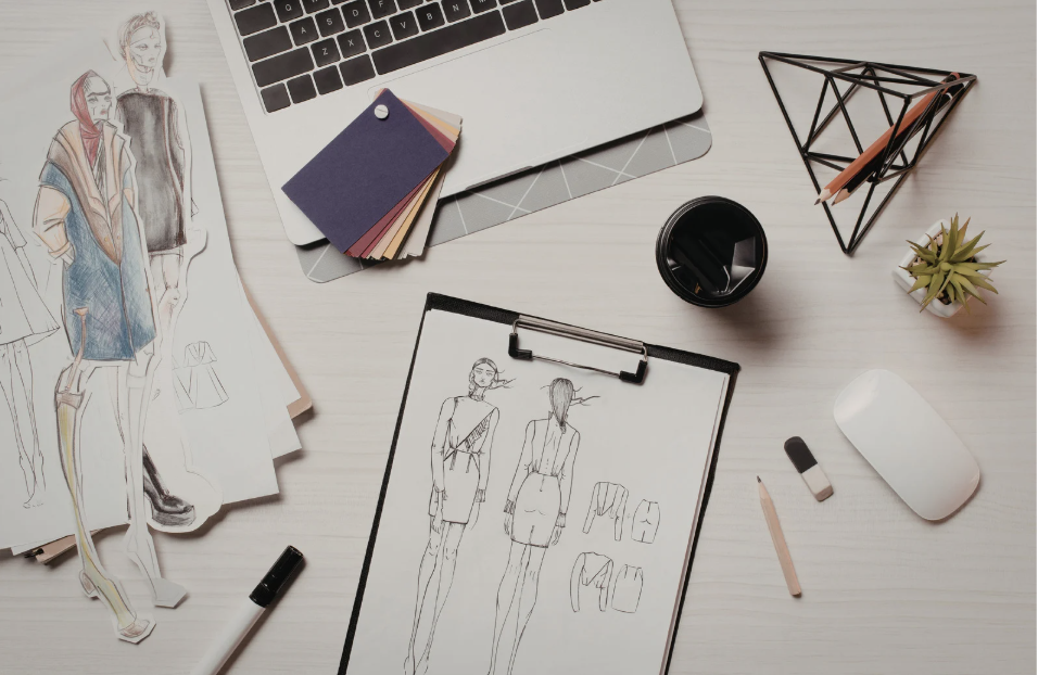 Fashion designer's workspace with sketches, fabric swatches, a laptop, drawing tools, and decorative items.