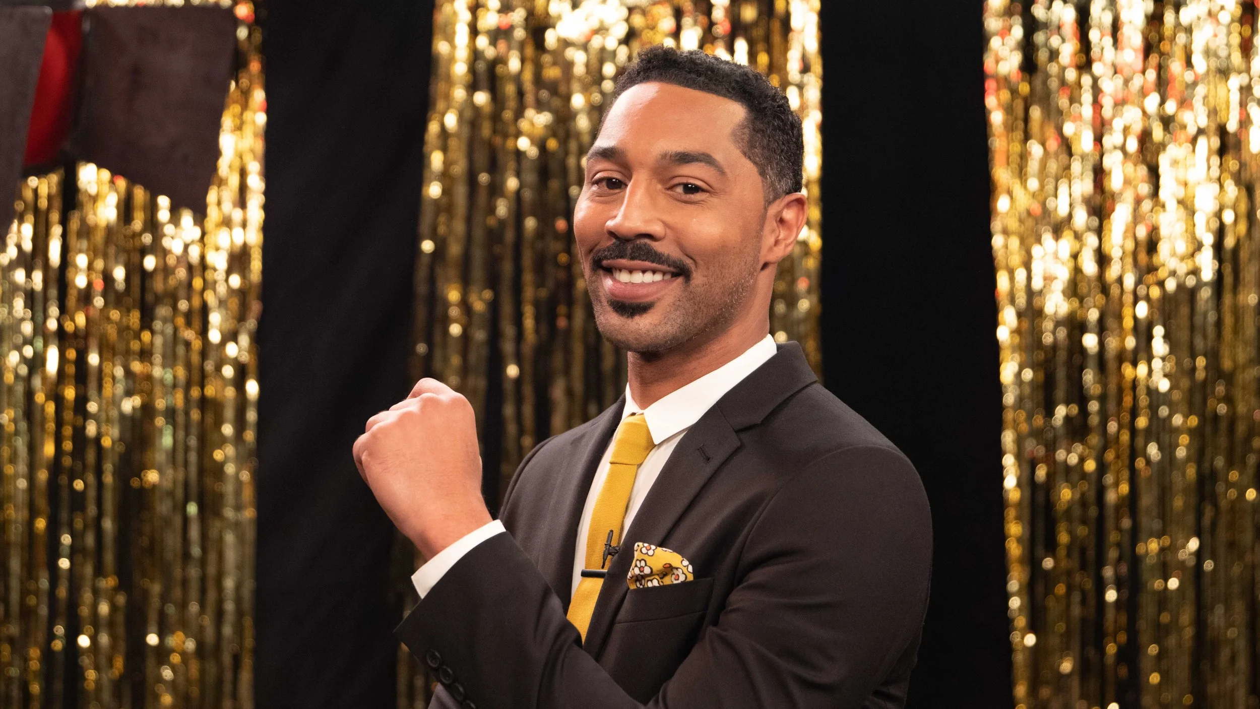 A smiling man in a black suit with a yellow tie and pocket square, posing with a flexed arm in front of a gold tinsel backdrop.