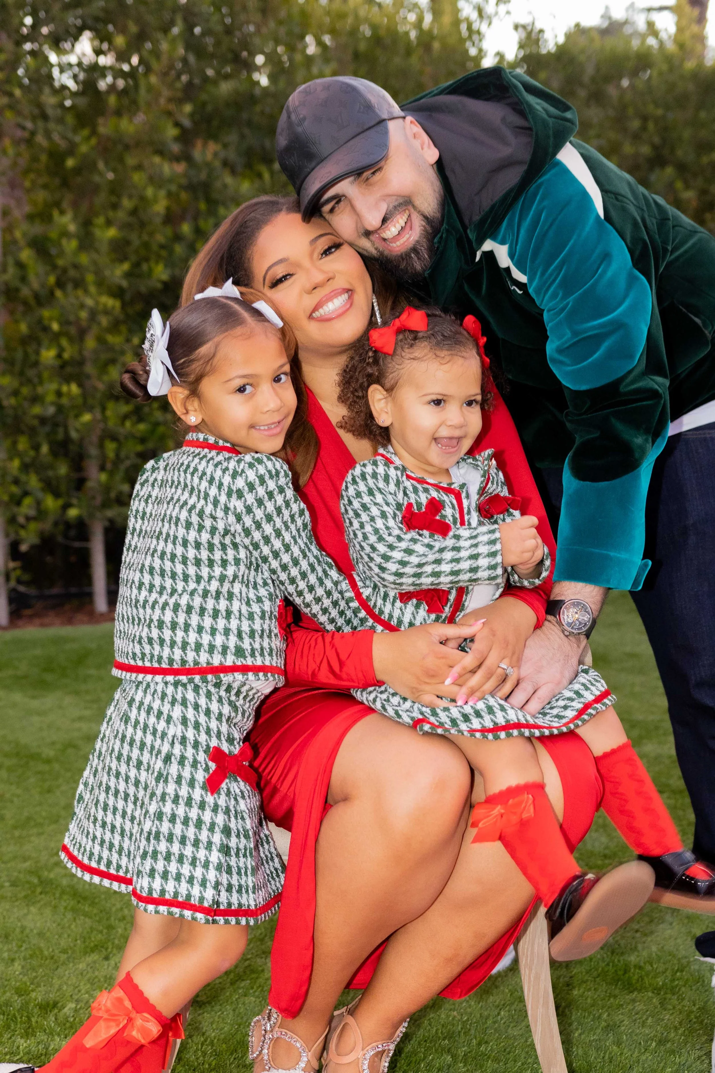 A family of four celebrating outdoors, smiling and dressed in festive holiday clothing, with two young girls in matching outfits, a woman in a red dress, and a man leaning over them, all posing happily.