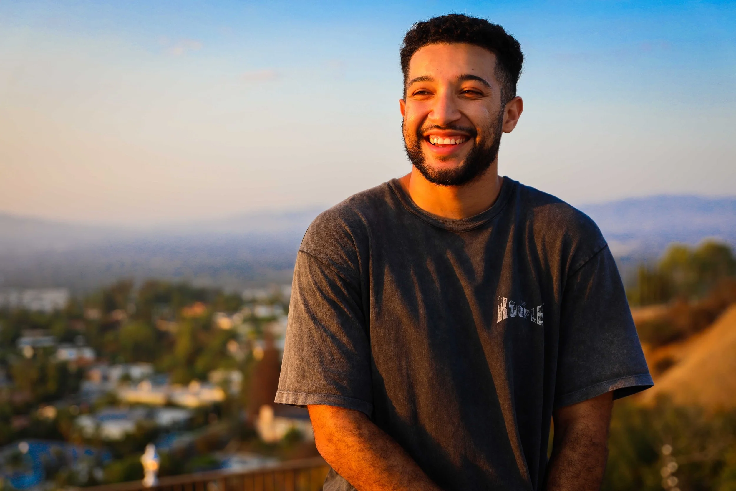 A smiling young man with dark hair and a beard standing outdoors during sunset, with a blurred cityscape and hills in the background.