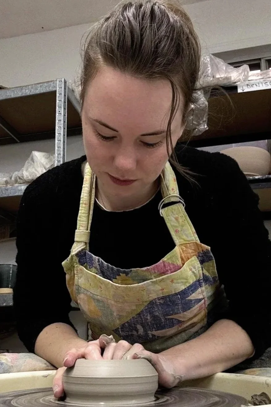 Young woman with brown hair in a bun wearing a colorful apron, focused on shaping clay on a pottery wheel in a studio.