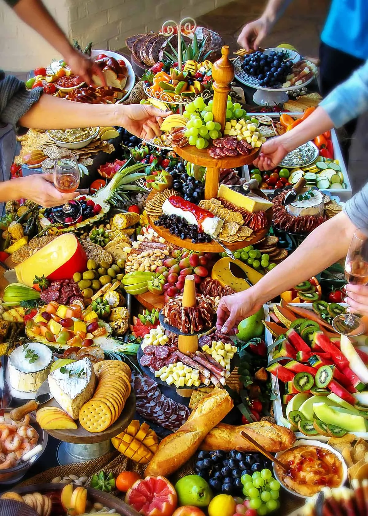 A large feast with various fruits, cheeses, meats, and desserts on a table, with people reaching for food.