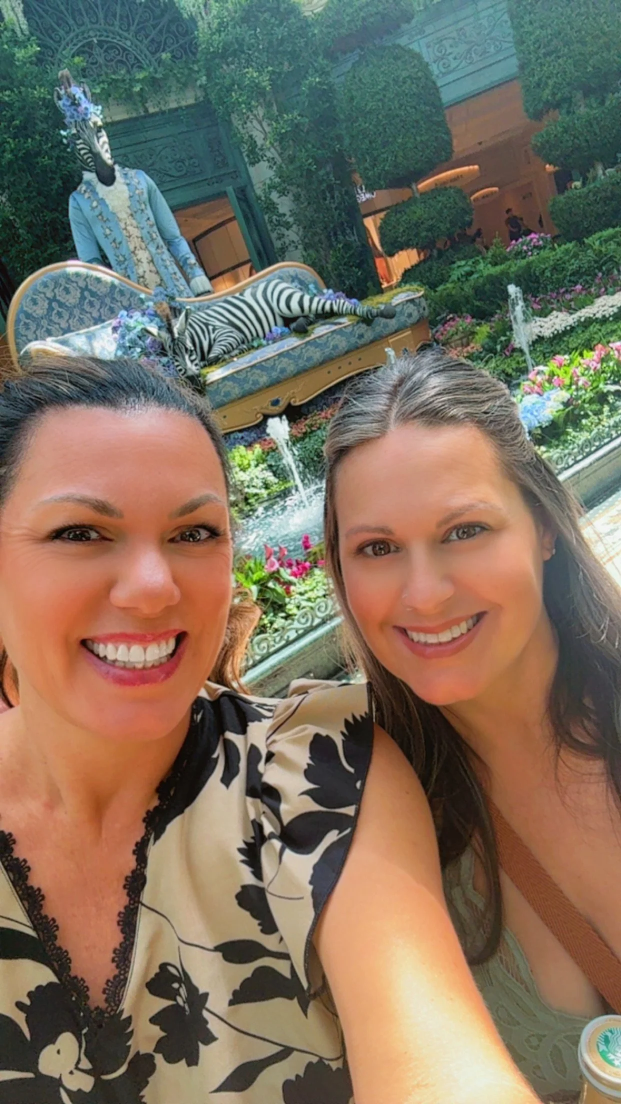 Two women smiling in front of a decorative indoor garden with a fountain, flowers, and a zebra in a glass display behind them.