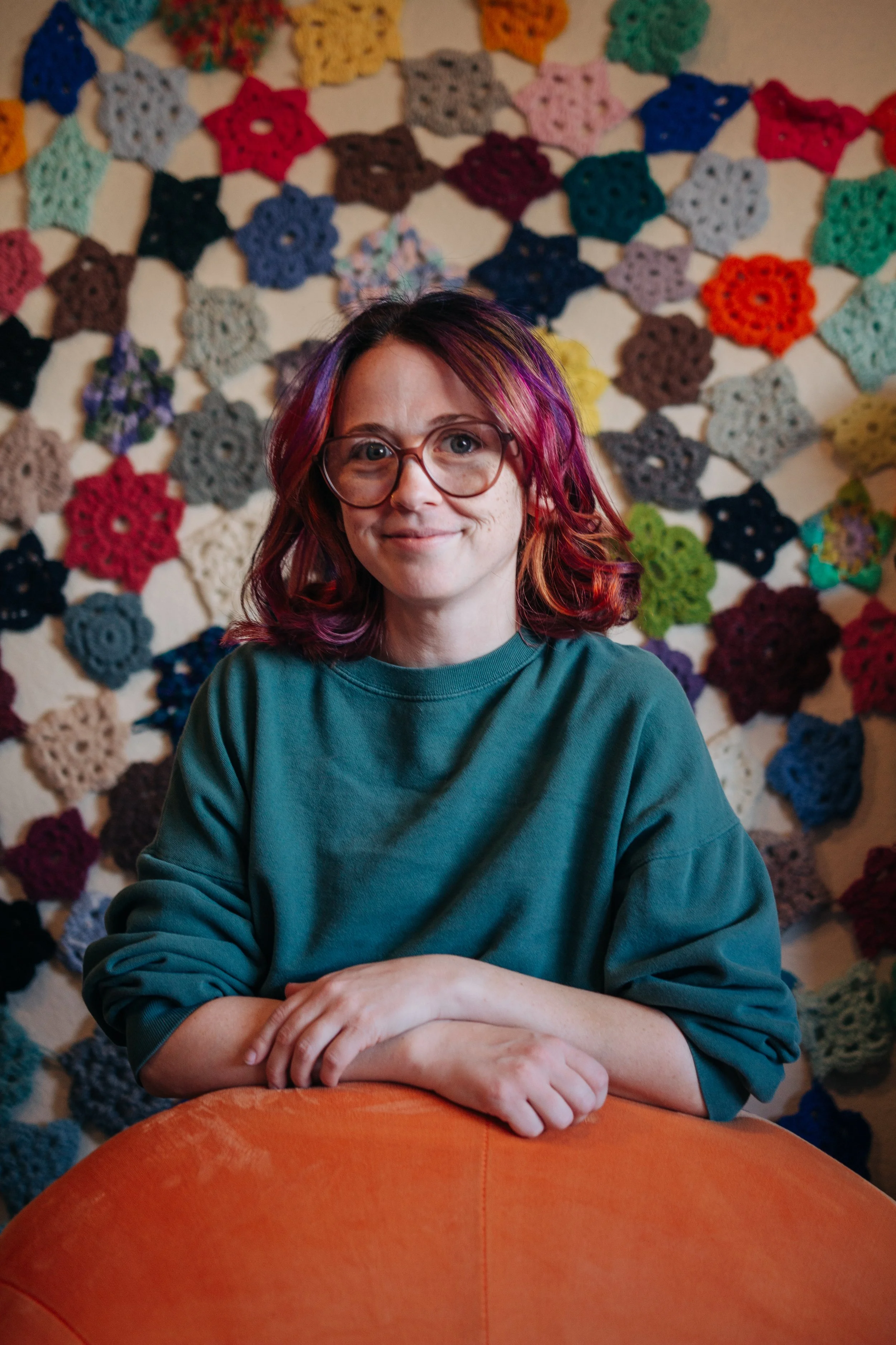 Art therapist Becca Meyers, with colorful hair, glasses, and a green sweater,leaning on the back of an orange chair with a crochet tapestry behind her