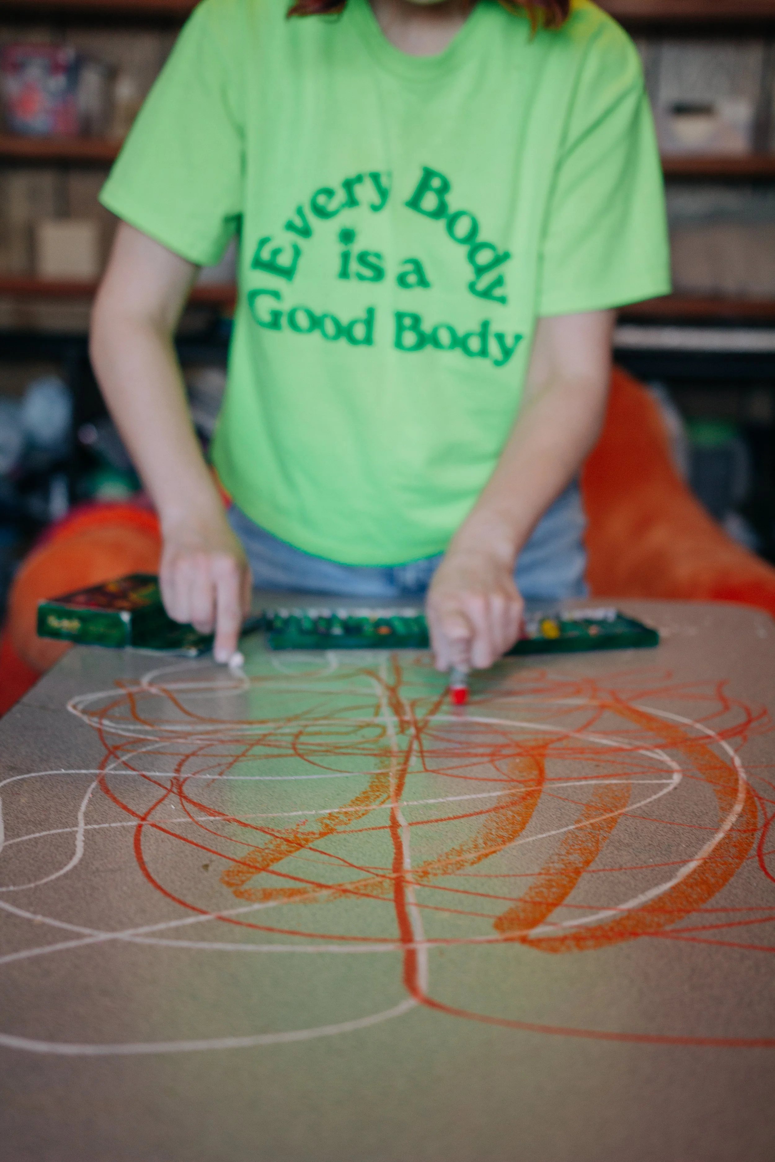 Becca Meyers wearing a shirt that says "every body is a good body" and using both hands to draw with oil pastels on a grey tabletop