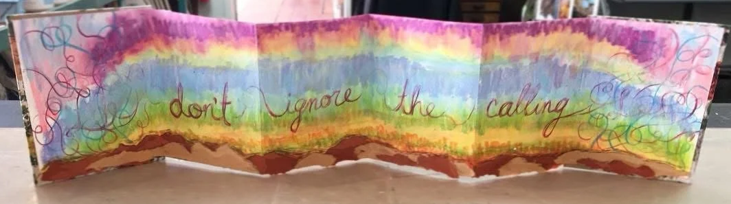 Accordian-style booklet filled with hand-drawn rainbow colors in horizontal lines, and torn paper across the bottom, featuring the handwritten message 'don't ignore the calling'.