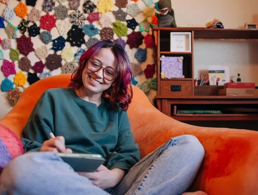 A woman with purple and pink hair, glasses, wearing a green sweatshirt and gray jeans, smiling and writing on a notepad while sitting on an orange couch in a cozy room decorated with a colorful crochet wall and wooden shelves.