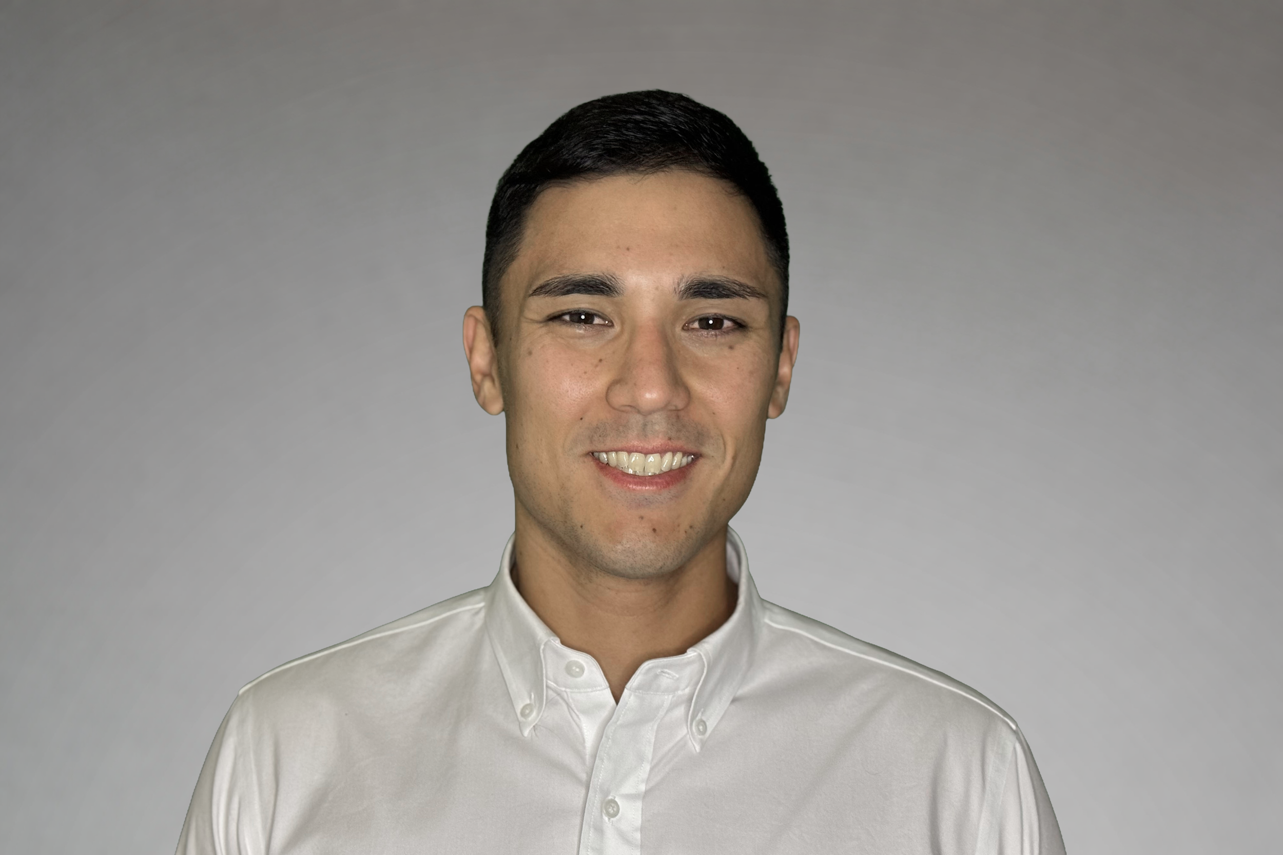 A young man with short dark hair, smiling, wearing a white button-up shirt, standing against a gray background.