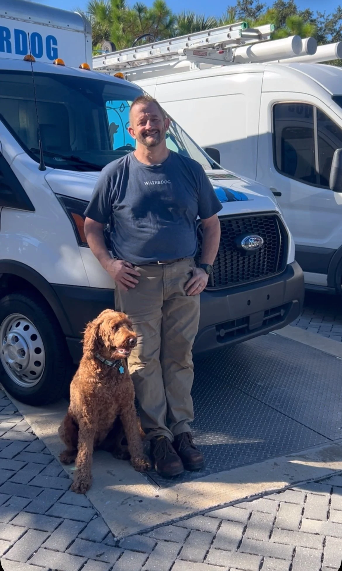 Man standing next to a happy brown dog in front of service vans with ladders on top, in a parking lot on a sunny day.