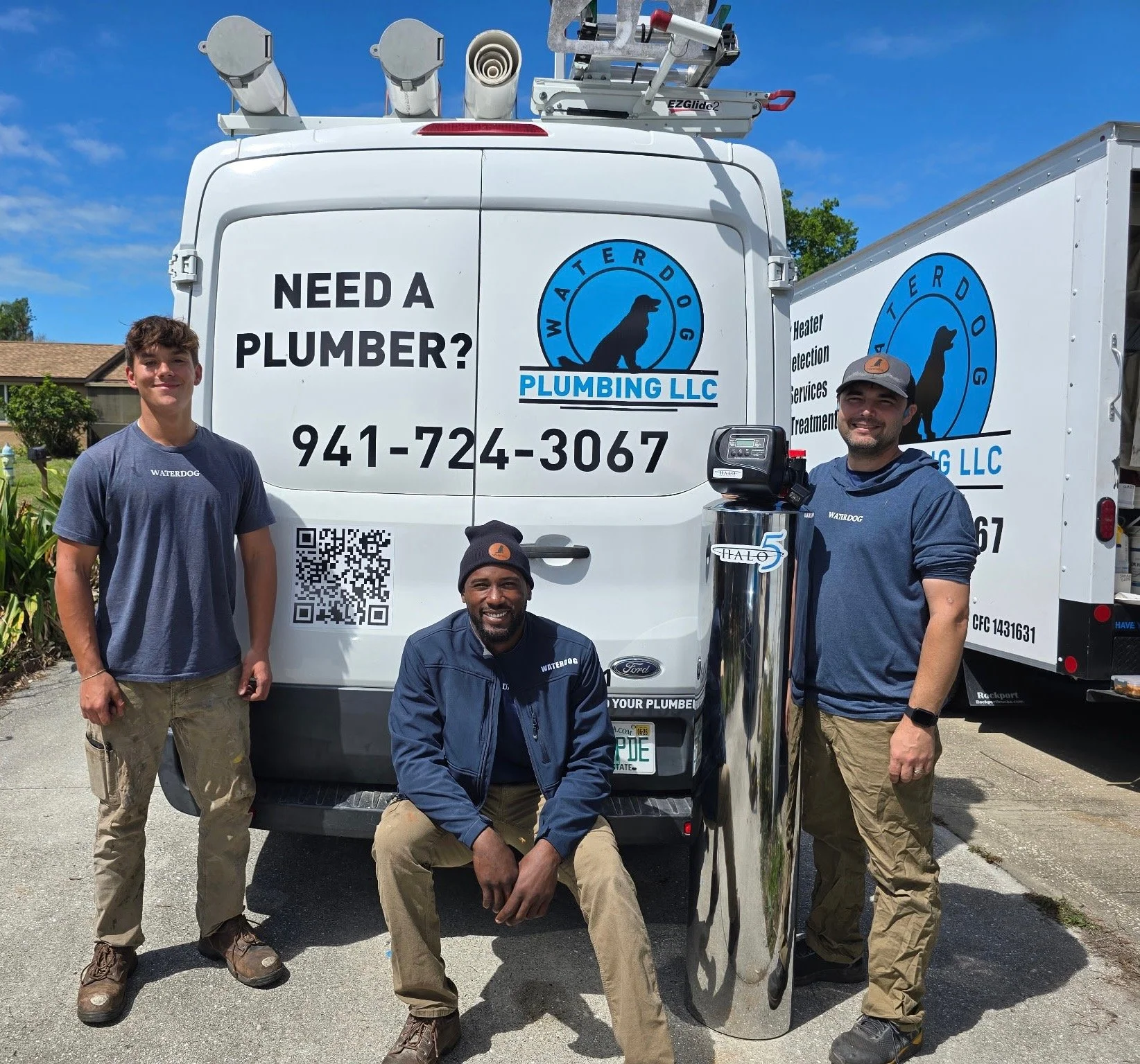 Three men standing and squatting in front of a white plumbing service van with the company logo and contact information. The men are wearing blue shirts and khaki pants, smiling at the camera on a sunny day.