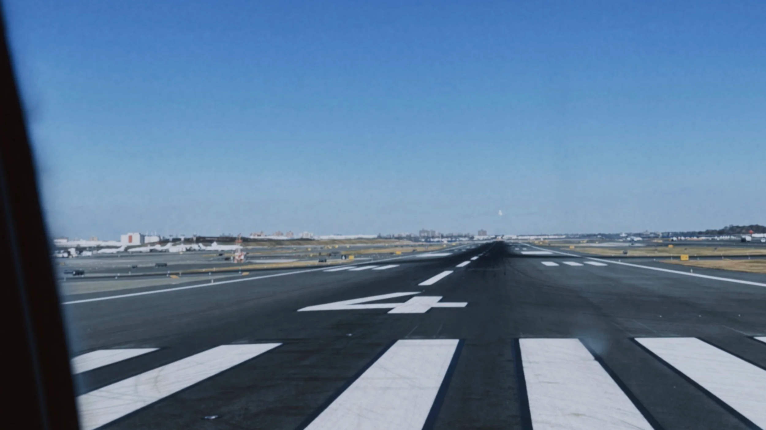 View of an airport runway seen from an aircraft window, with runway markings and airport buildings in the background under a clear blue sky.