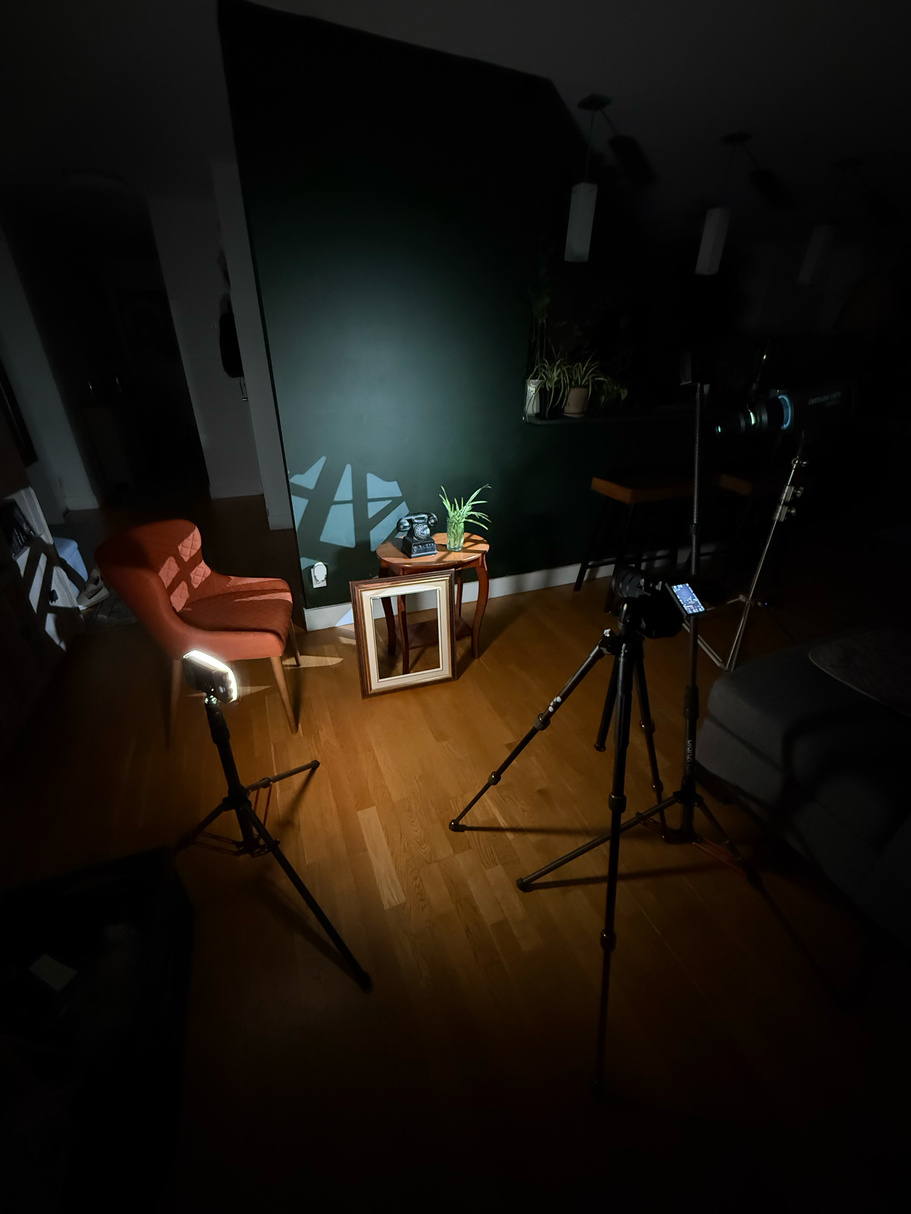 Living room with a green accent wall, orange armchair, side table with a vintage telephone and potted plant, and a photography or film setup with a camera and lighting equipment.