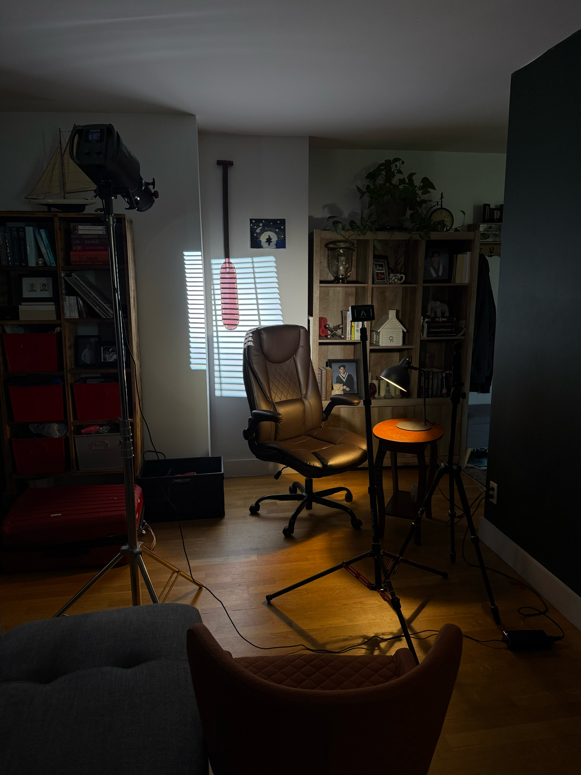 A dimly lit room with a brown leather office chair, wooden bookshelf filled with books and decor, a small table with a lamp, a potted plant, and photography or filming equipment including a light stand and tripod.