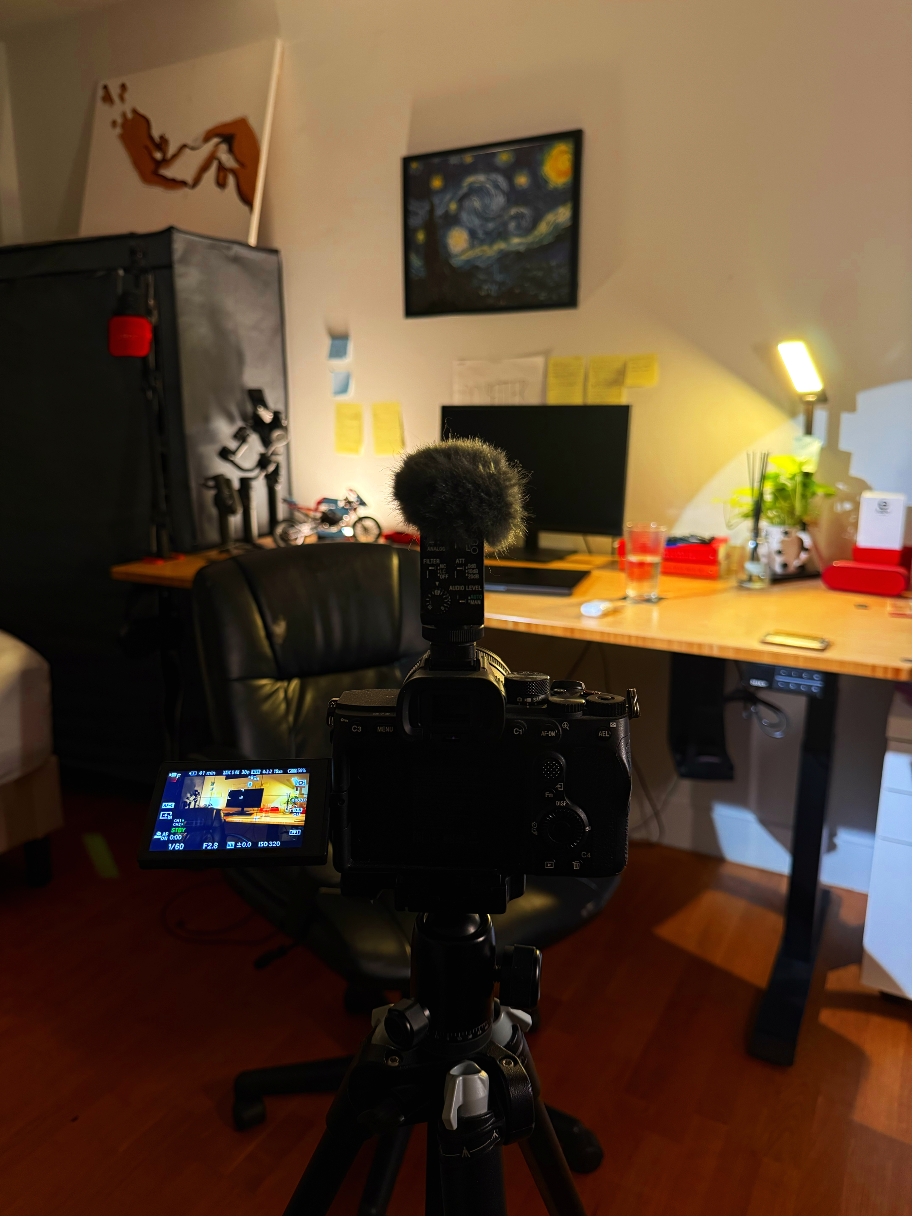 An indoor workspace with a camera on tripod aimed at a desk. The desk has a computer monitor, a backlit lamp, a glass of water, a plant, and various objects. There are sticky notes on the wall, a model bicycle, and artwork including Van Gogh's Starry Night.