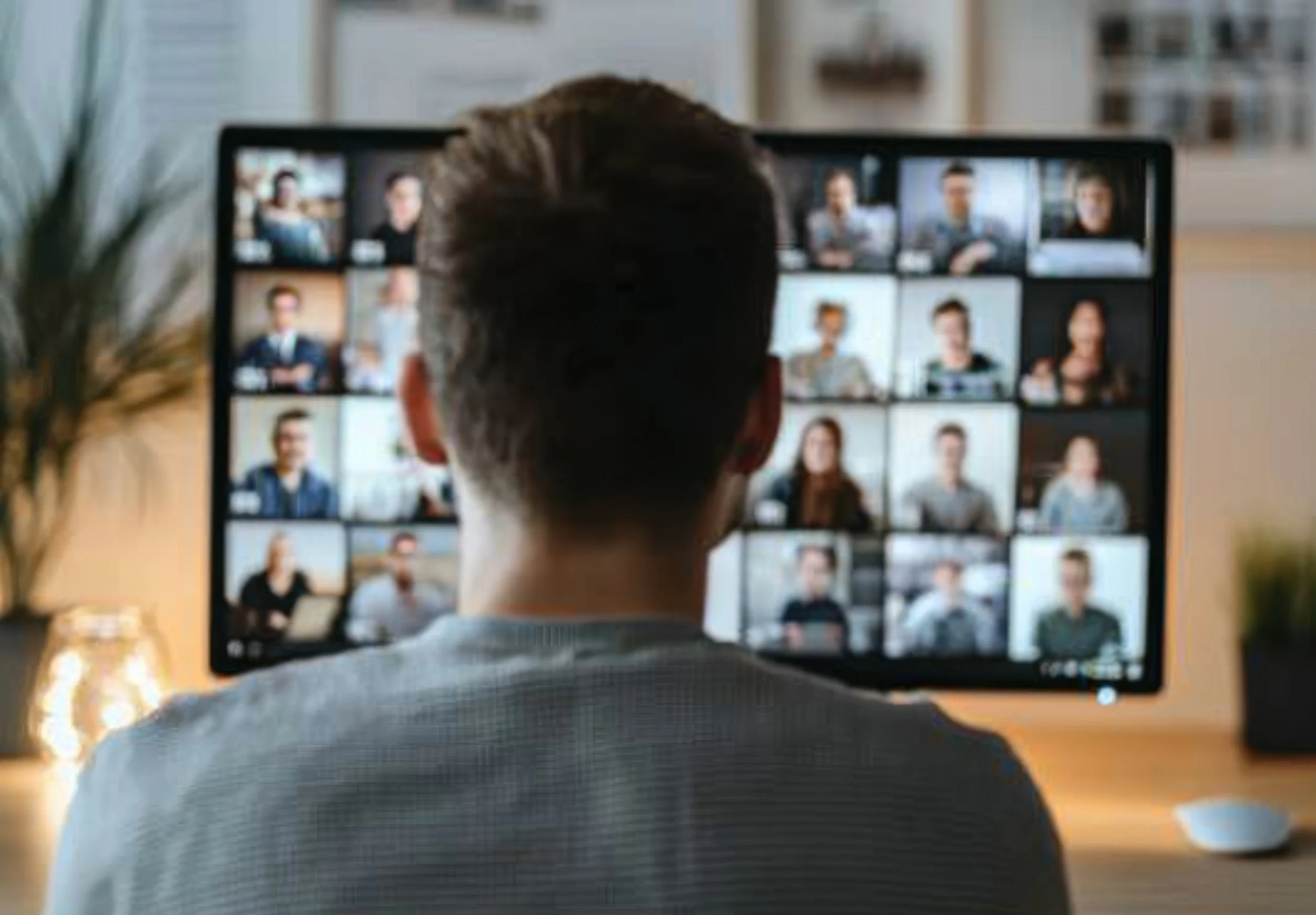 Person with short hair is sitting at a desk, participating in a video conference with multiple people displayed on a large monitor in a grid layout.