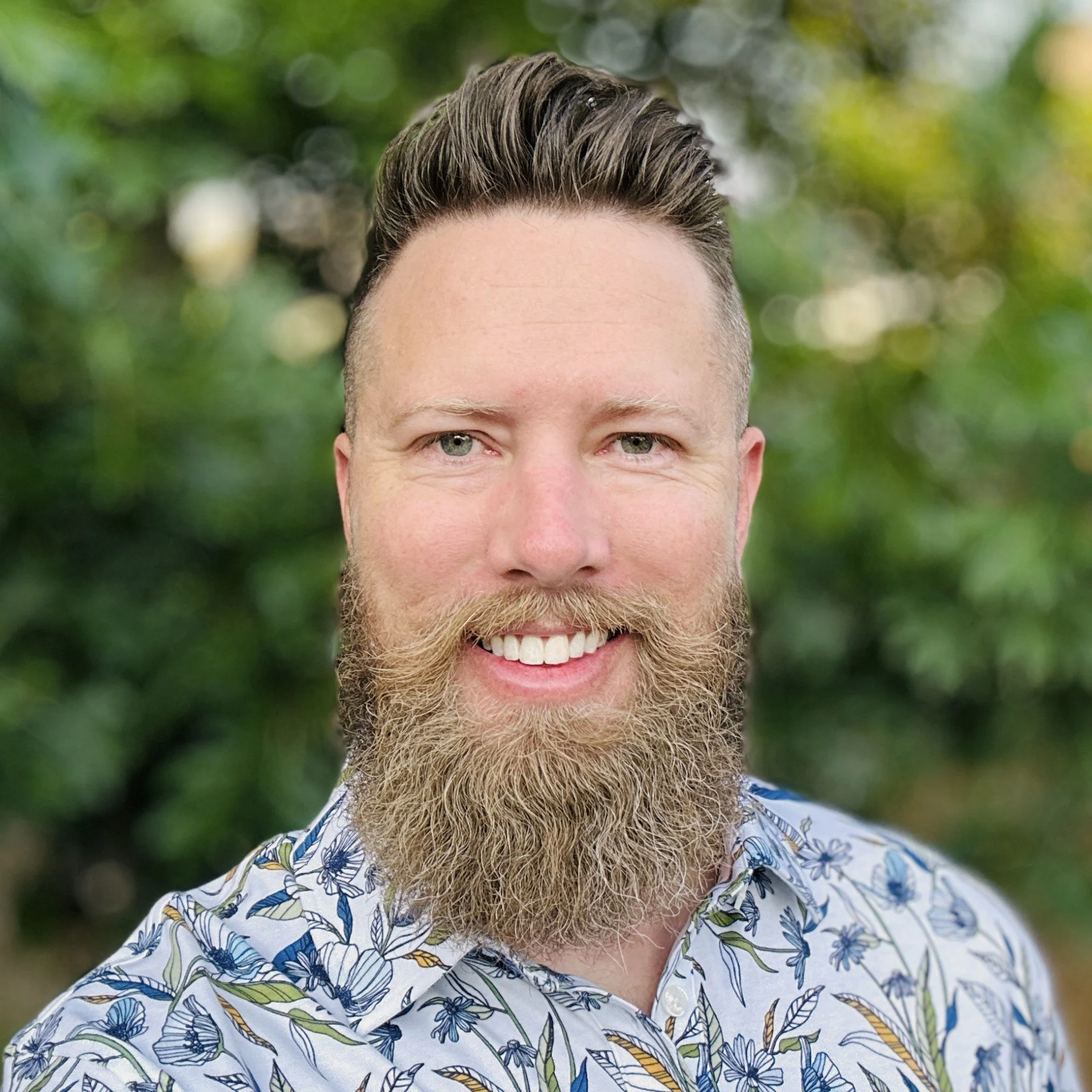 A man with a full beard and styled hair smiling outdoors with a blurred green leafy background, wearing a button-up shirt.