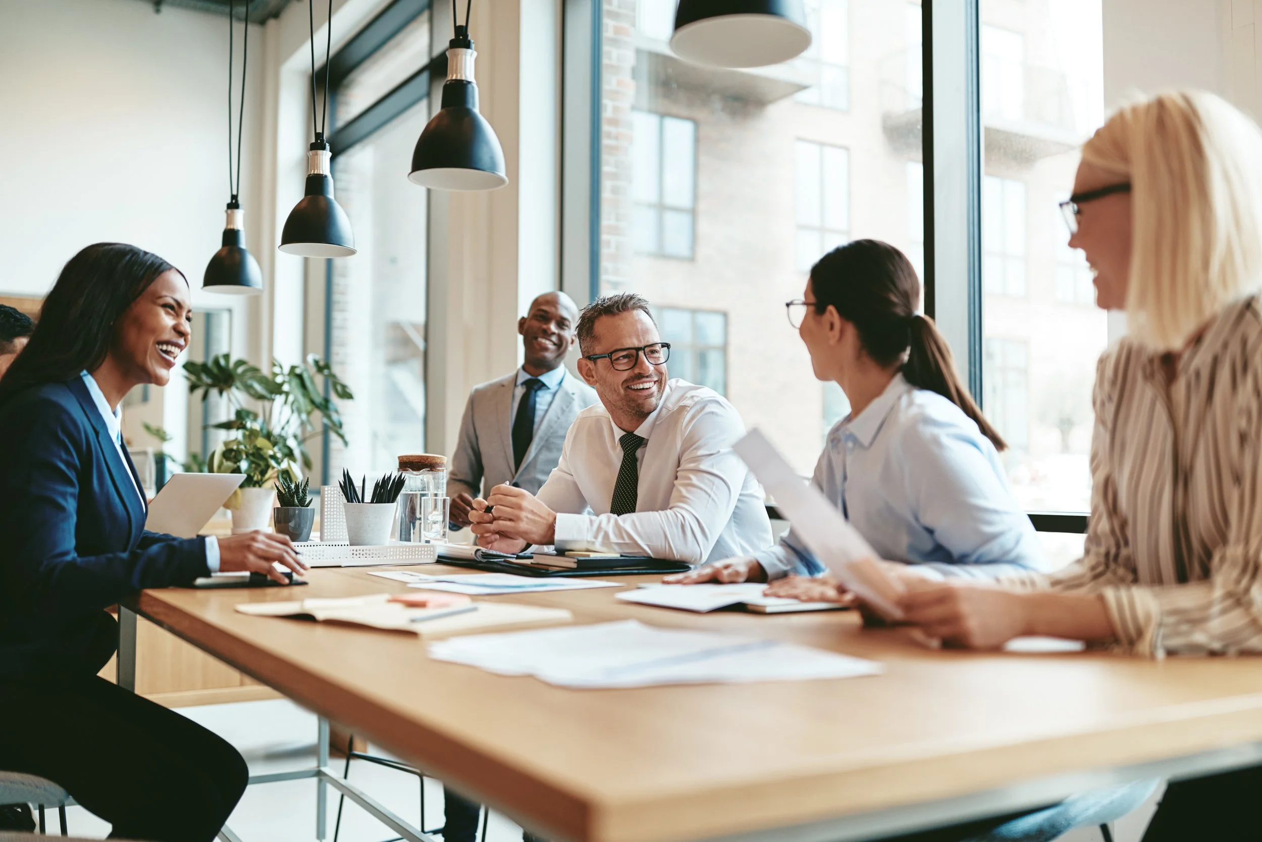 Business meeting with five diverse professionals sitting at a table, smiling and engaging in conversation, in a bright modern office with large windows and hanging pendant lights.