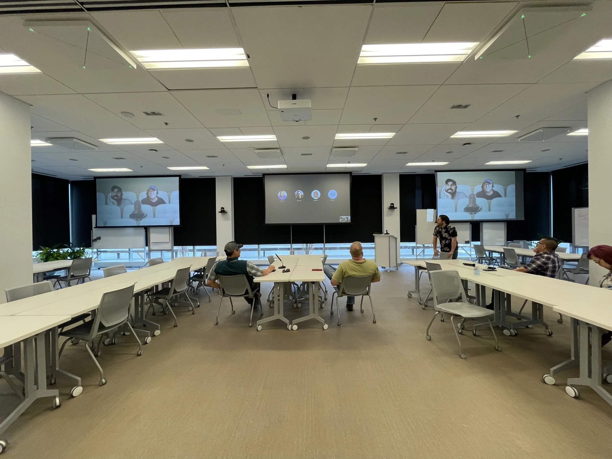 A conference room with people seated at tables, a presenter standing, and multiple large screens displaying the audience.