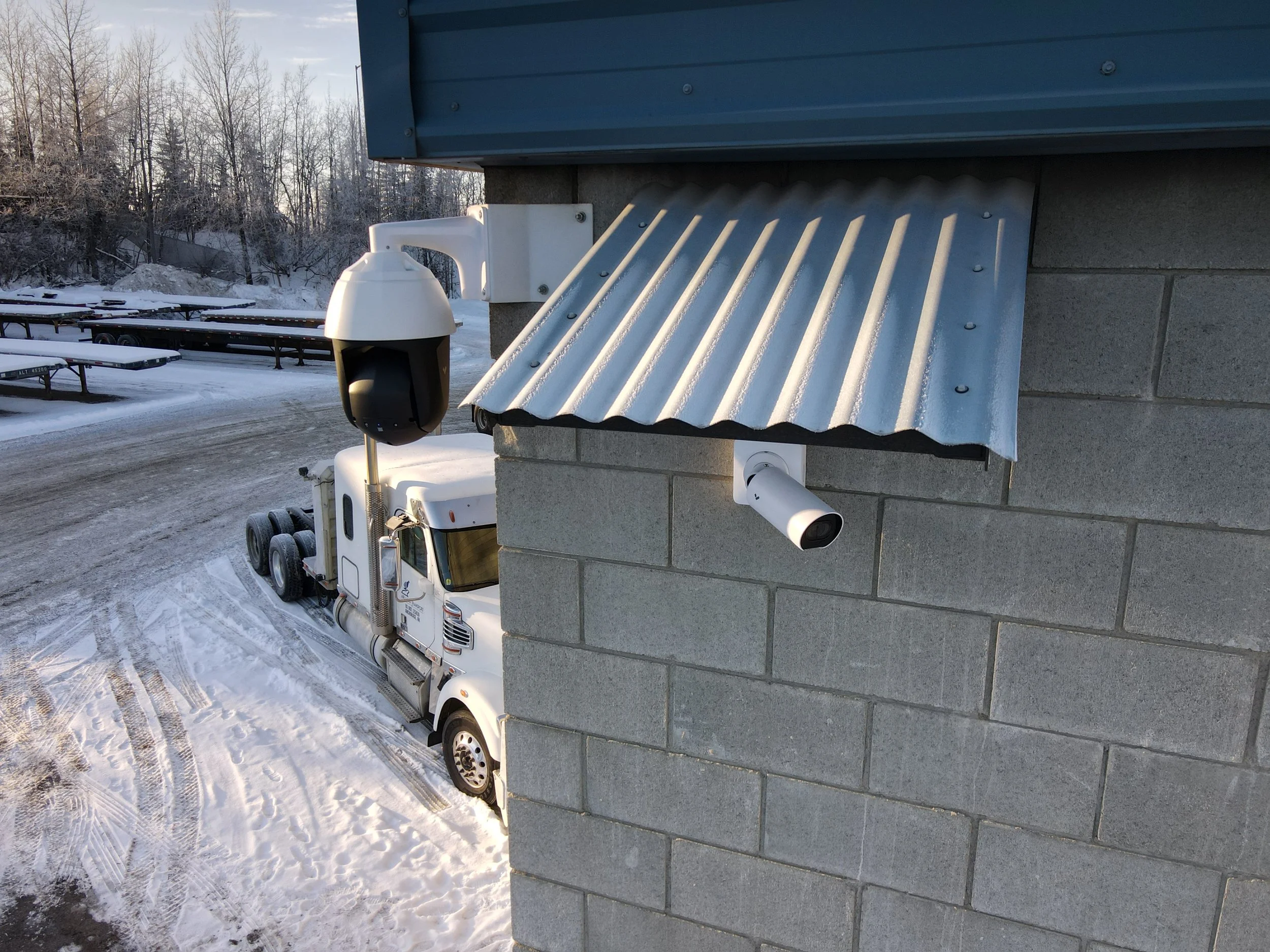 Surveillance camera mounted on a brick wall outside in snowy weather, with a truck parked below.