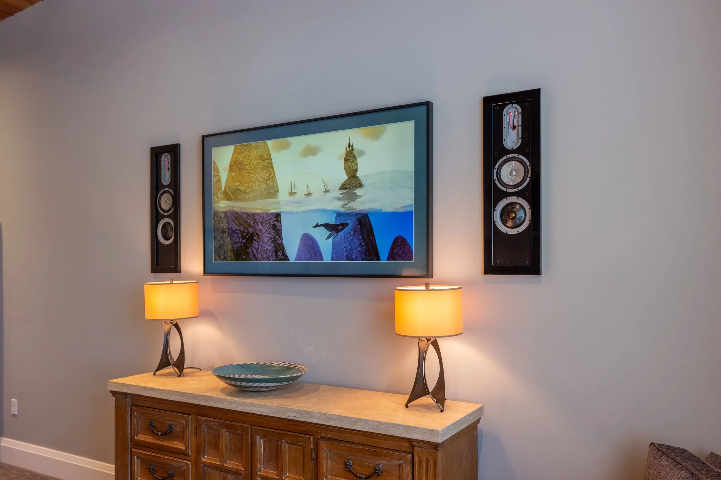 Living room wall with a framed animated underwater scene painting, two black vertical speakers on either side, a wooden console table with a decorative bowl, and two matching table lamps with beige shades.