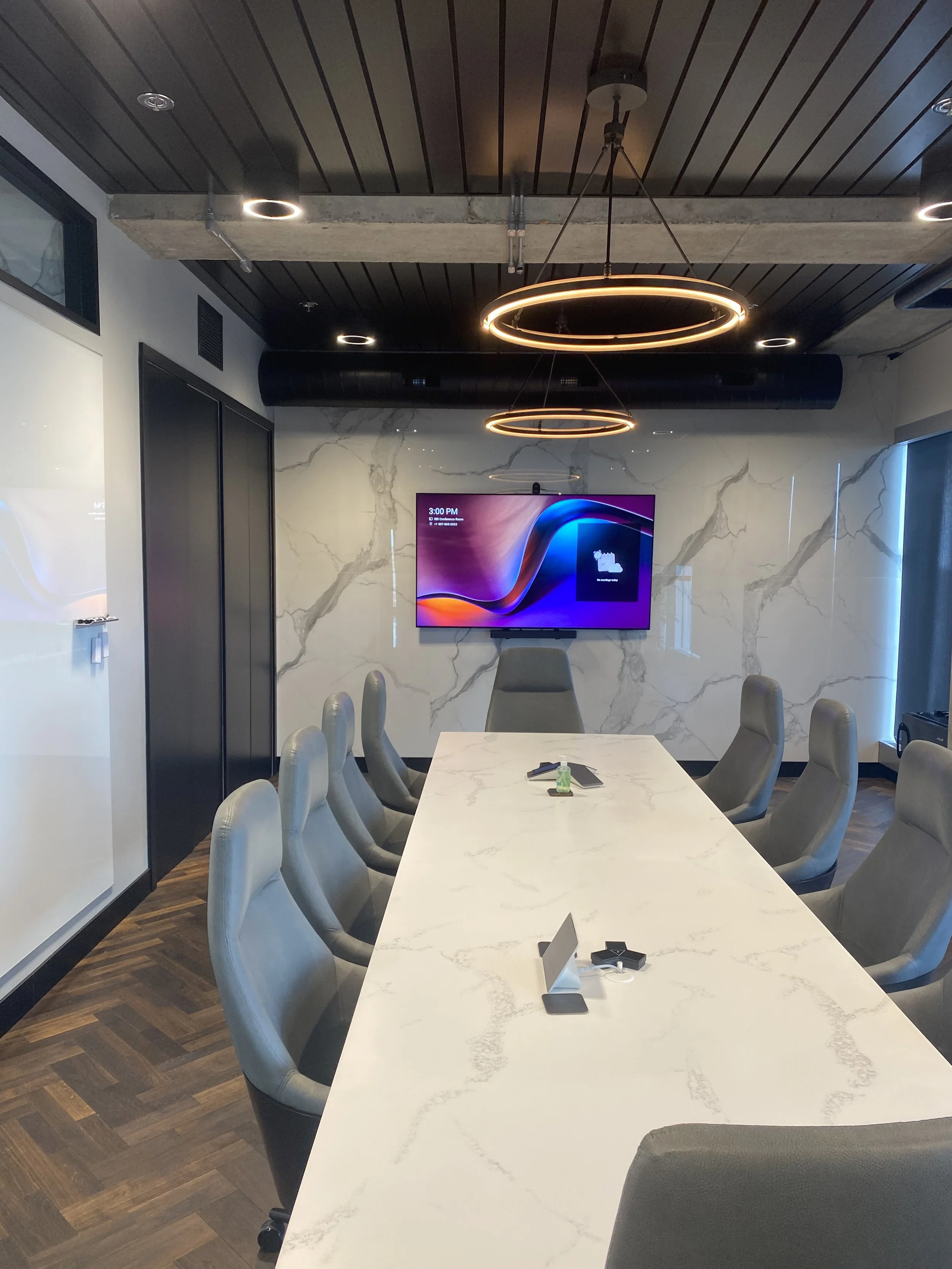Modern conference room with white marble table, gray chairs, a large wall-mounted screen, marble wall, and contemporary lighting fixtures.