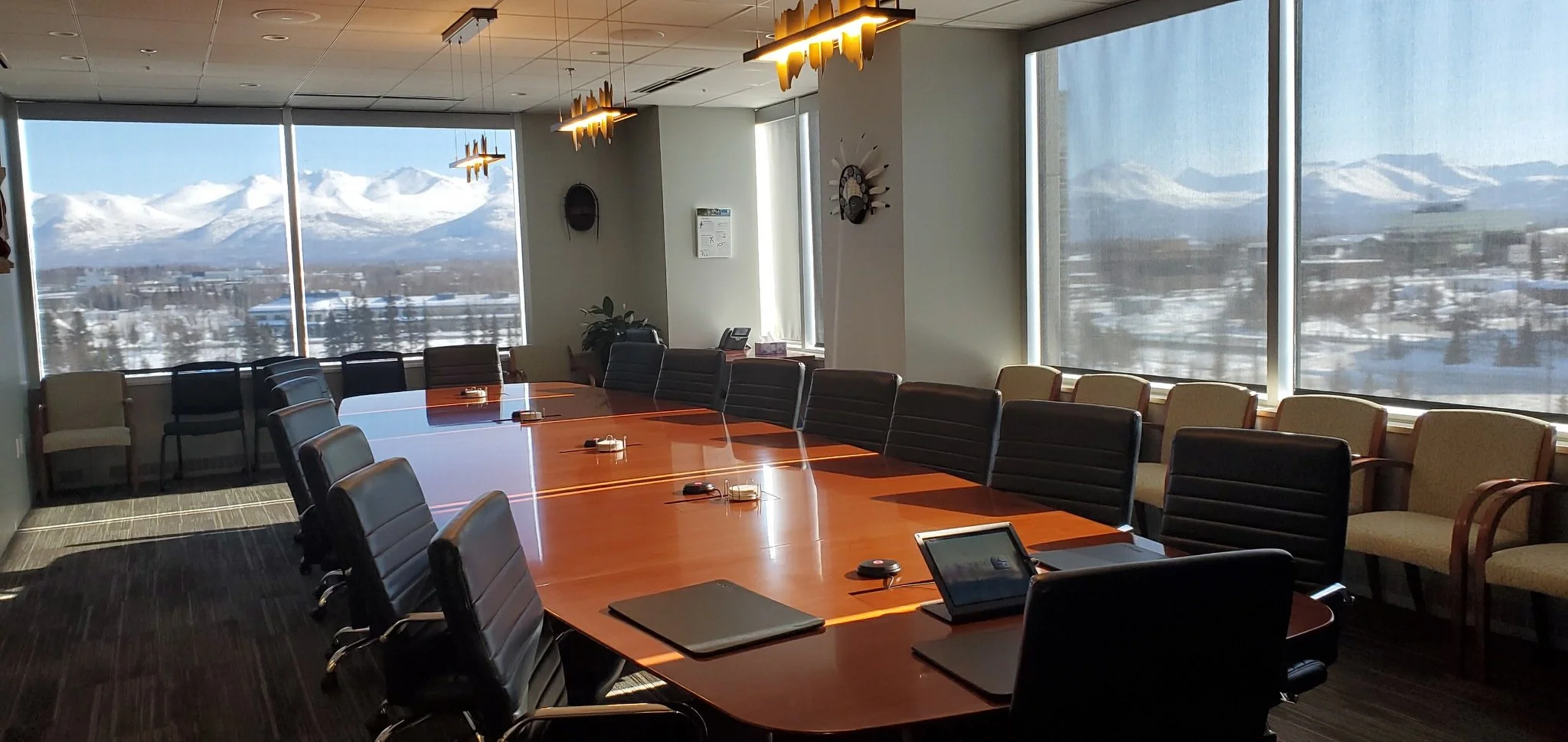 A conference room with a large wooden table and black leather chairs, large windows showing snow-covered mountains and landscape, with modern light fixtures and a few office items on the table.