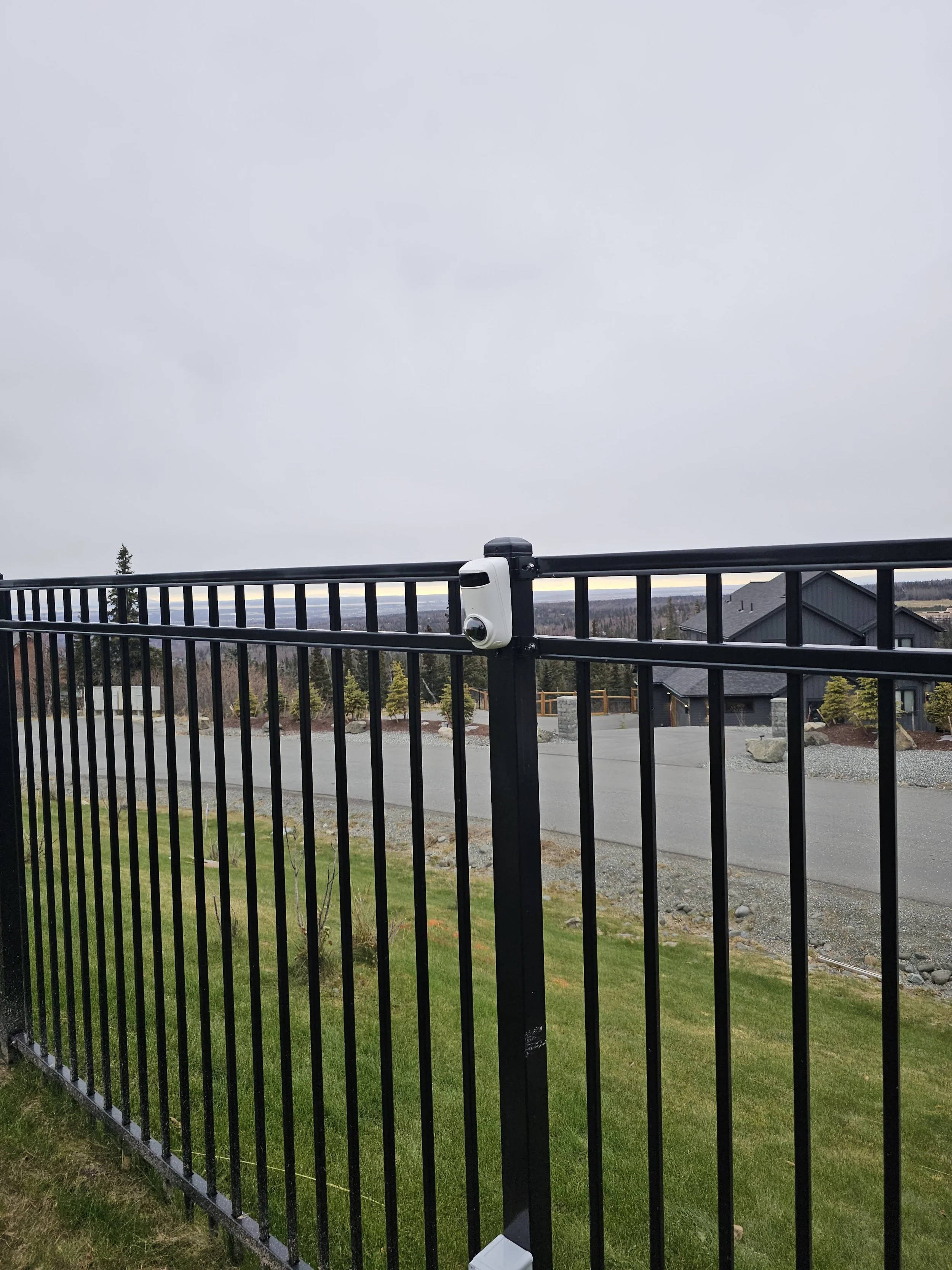 Black metal fence with a white security camera mounted on a post in a suburban yard. Overcast sky in the background.