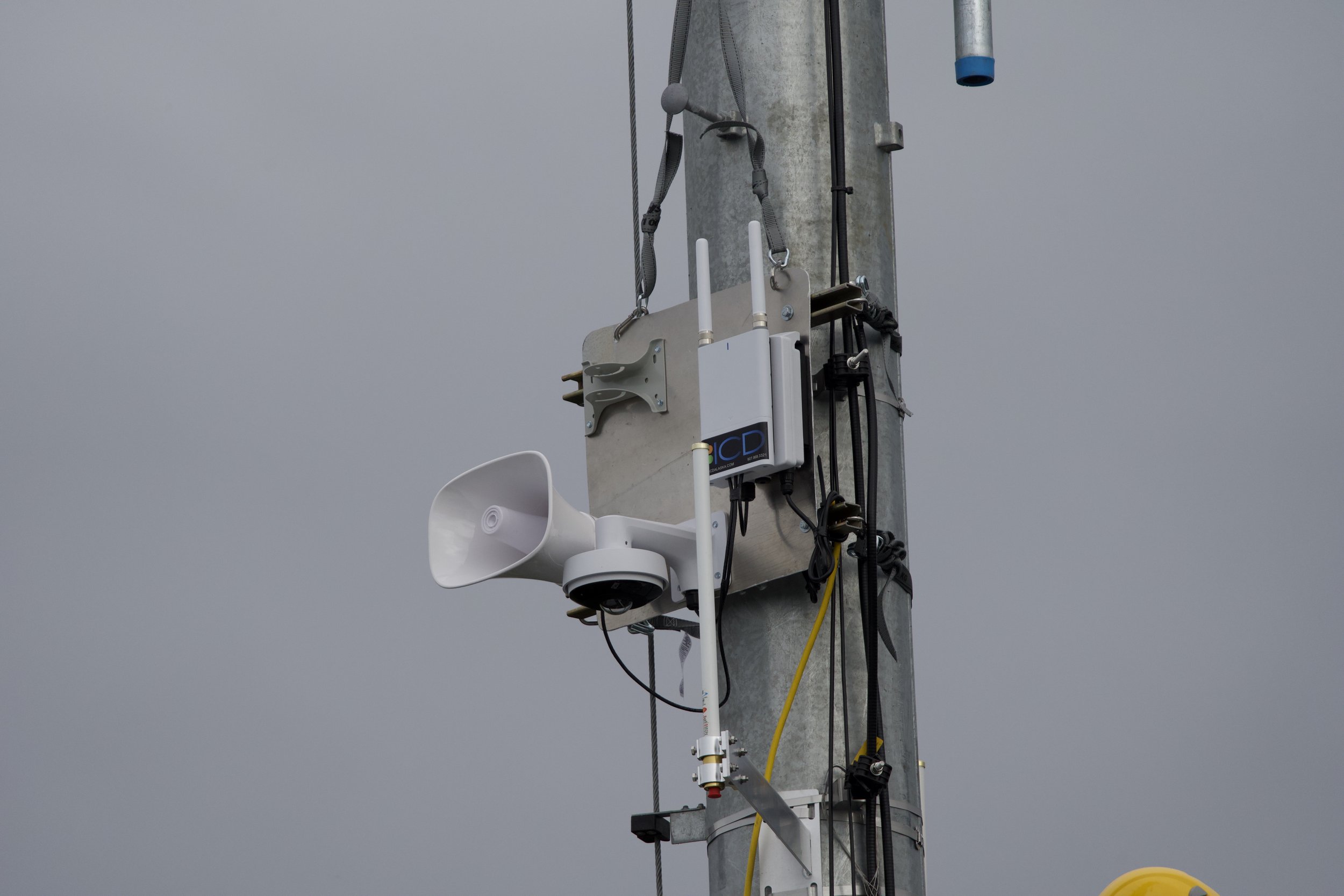 Verkada communication and video surveillance equipment attached to a metal utility pole, including a loudspeaker, antennas, and a 360º camera, against a cloudy sky.