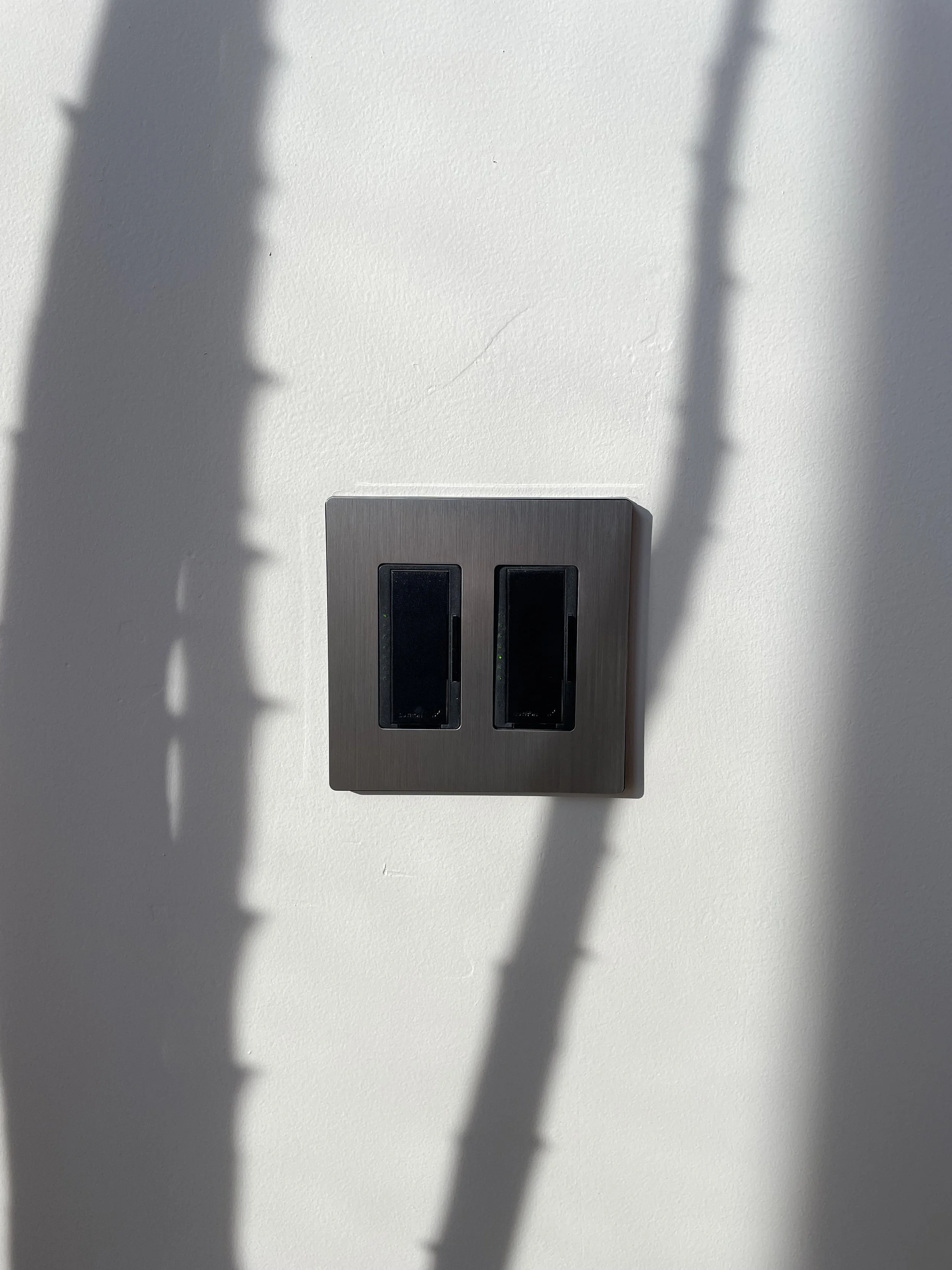 Lutron dimmers in midnight color in a 2-gang stainless steel faceplate a white wall with shadows cast across the wall.