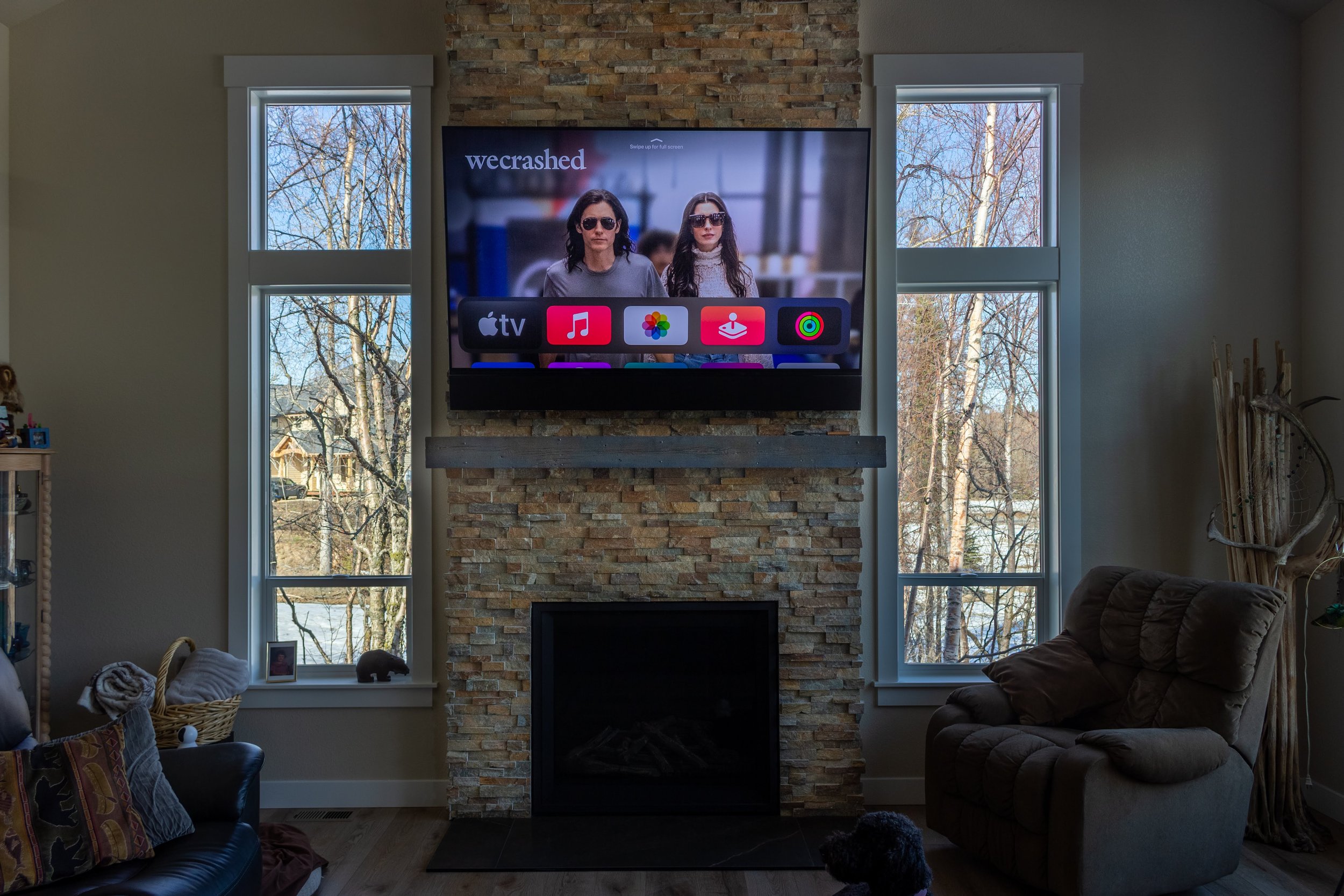 Living room with a stone fireplace, two large windows showing leafless trees outside, and a mantle-mounted Sony TV displaying a scene from the show 'WeCrashed' with two women wearing sunglasses, along with app icons below the scene.