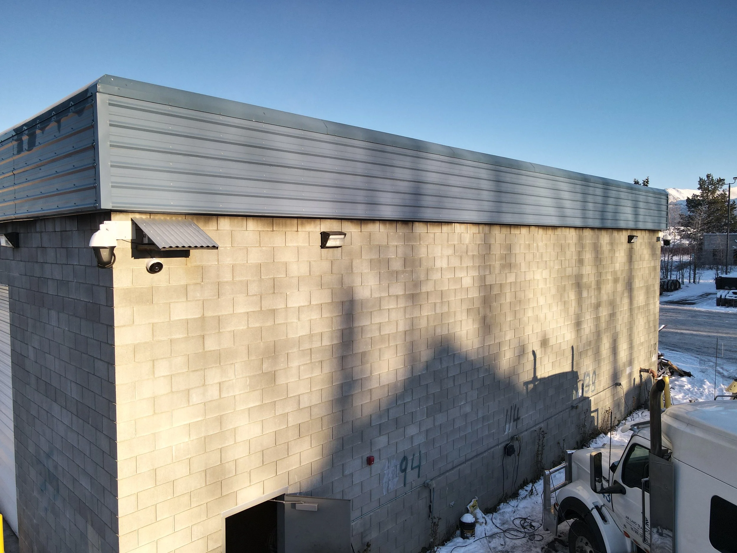 Exterior view of a warehouse or industrial building with a beige brick wall, a metal roof, Verkada security cameras, and outdoor lighting, with snow on the ground and a snow-covered mountain in the background.